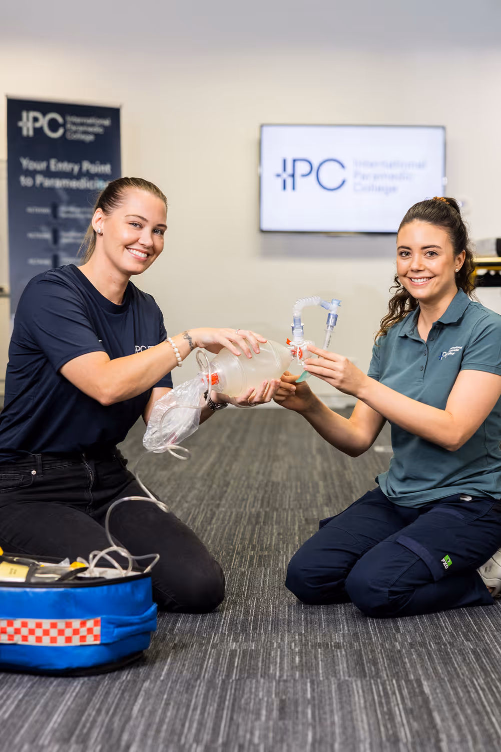 Two young women smiling and kneeling on a carpeted floor, demonstrating use of a manual resuscitator bag in a paramedic training setting.