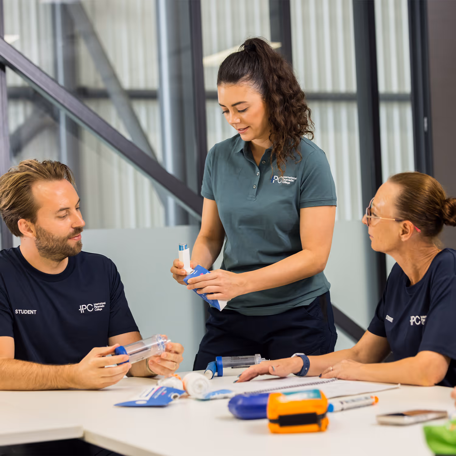 Three paramedic students in a classroom handling medical training devices including inhalers and airway equipment.