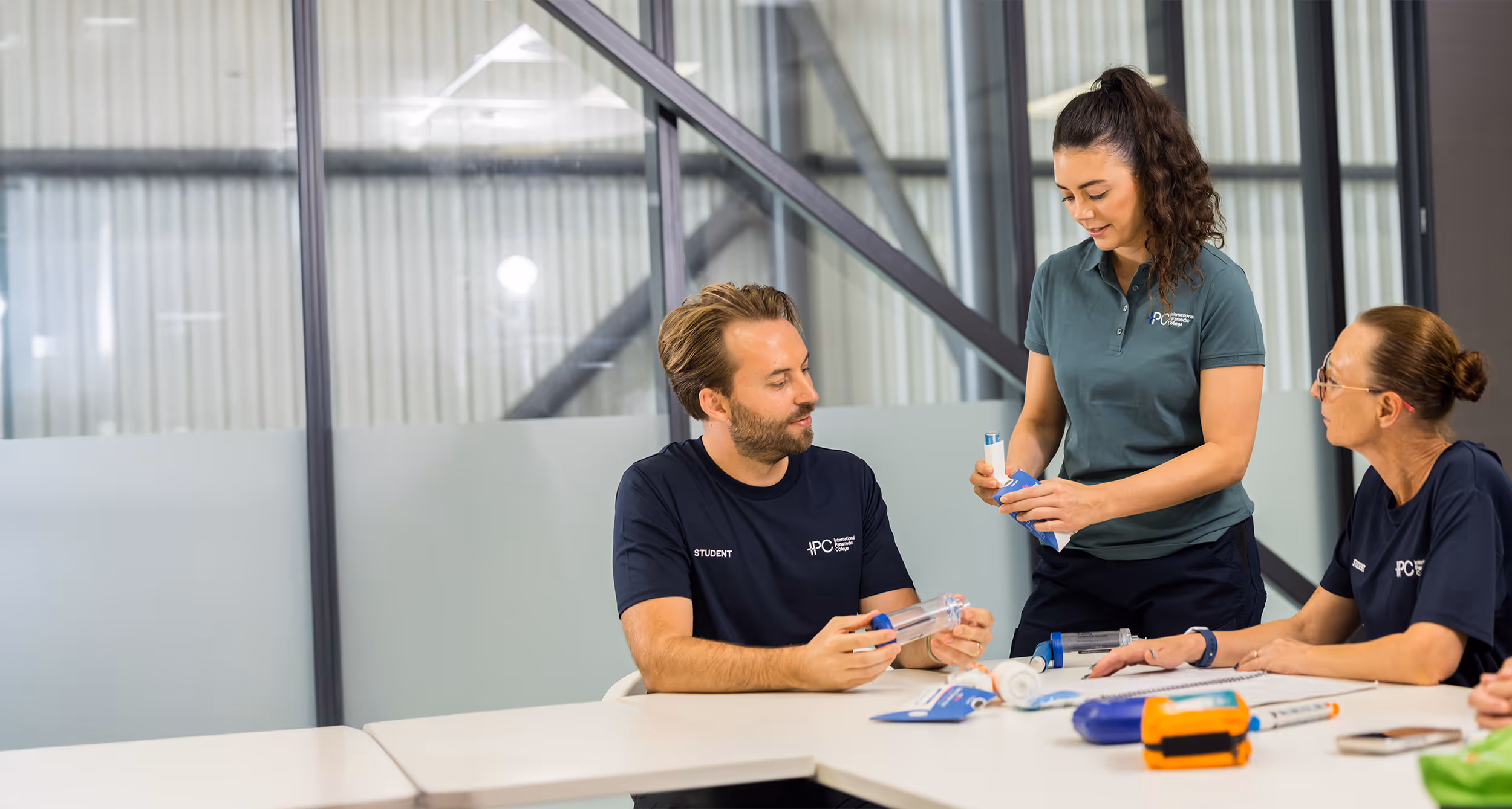 Three people in IPC International Paramedic College uniforms practicing with medical training devices at a classroom table.