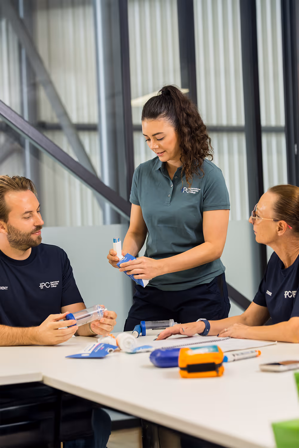 Three paramedic students in a classroom setting examining medical training equipment on a table.