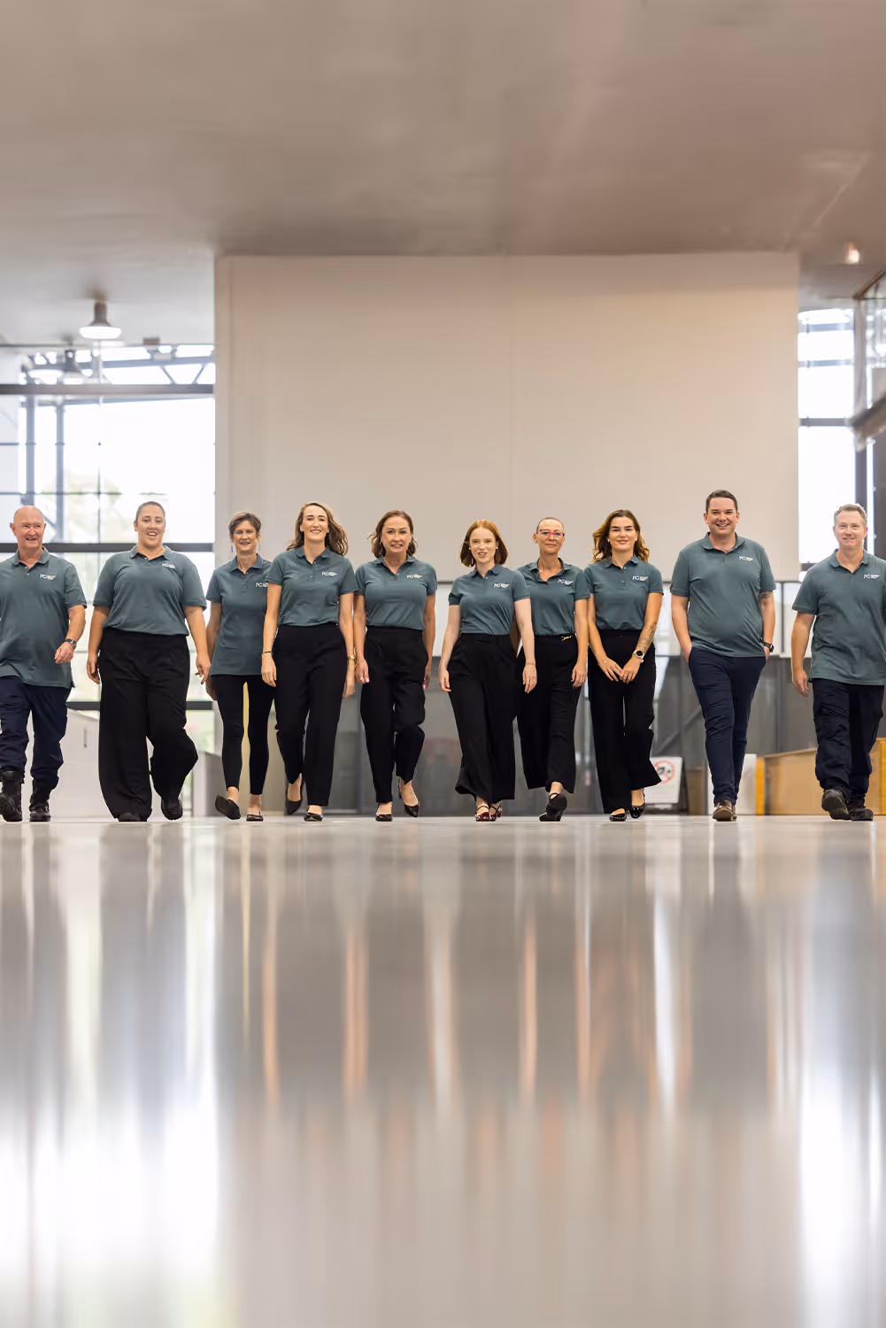 Group of ten people walking confidently indoors wearing matching teal polo shirts and black pants.