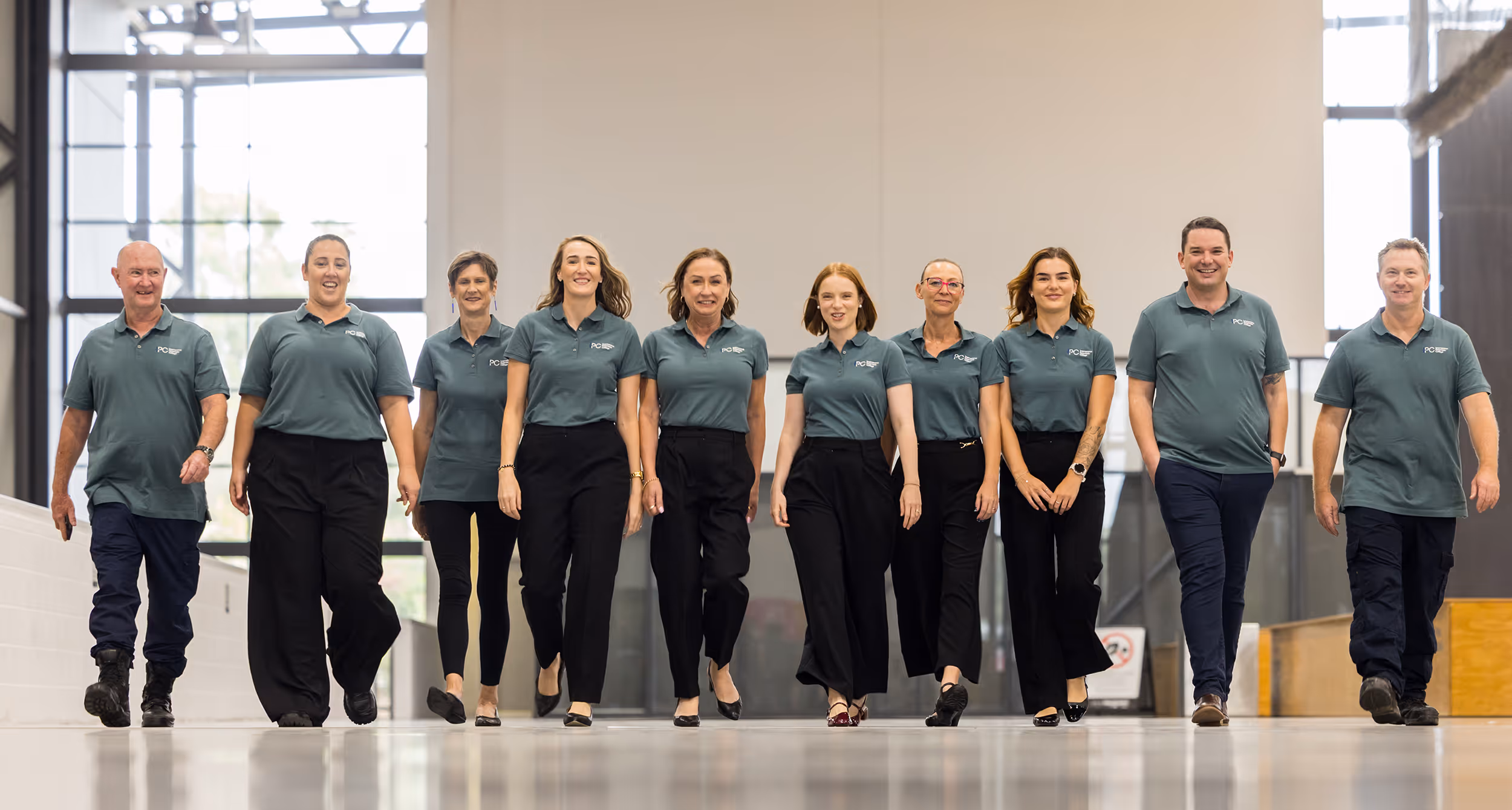 Group of ten smiling people walking forward indoors wearing matching green polo shirts and black pants.