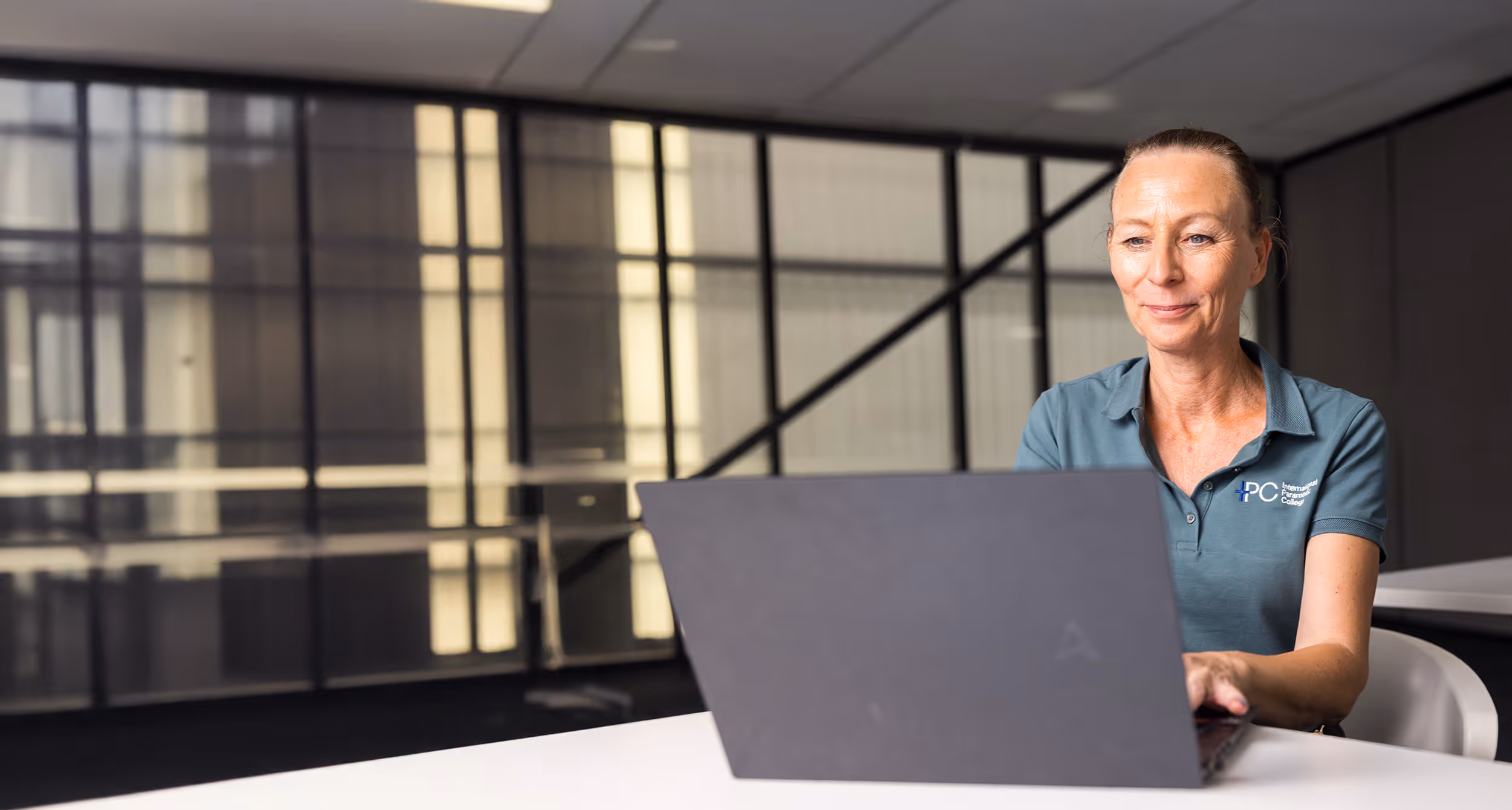 Woman in a teal polo shirt with IPC logo working on a laptop in a modern office setting.