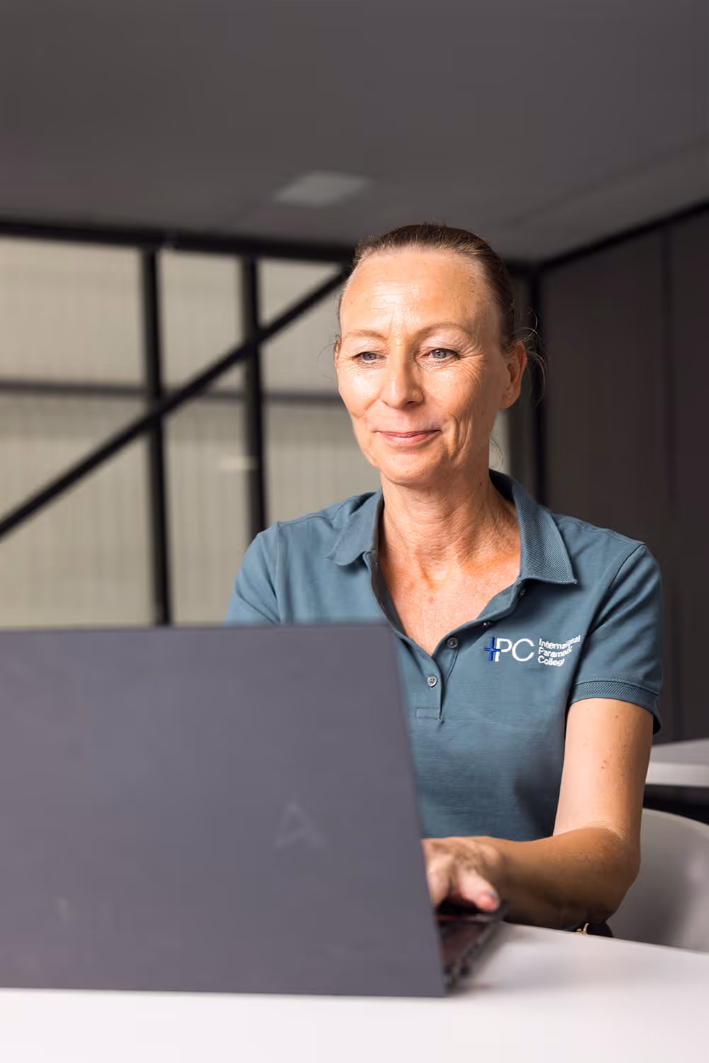 Woman in a teal polo shirt with International Paramedic College logo working on a laptop in a modern office.