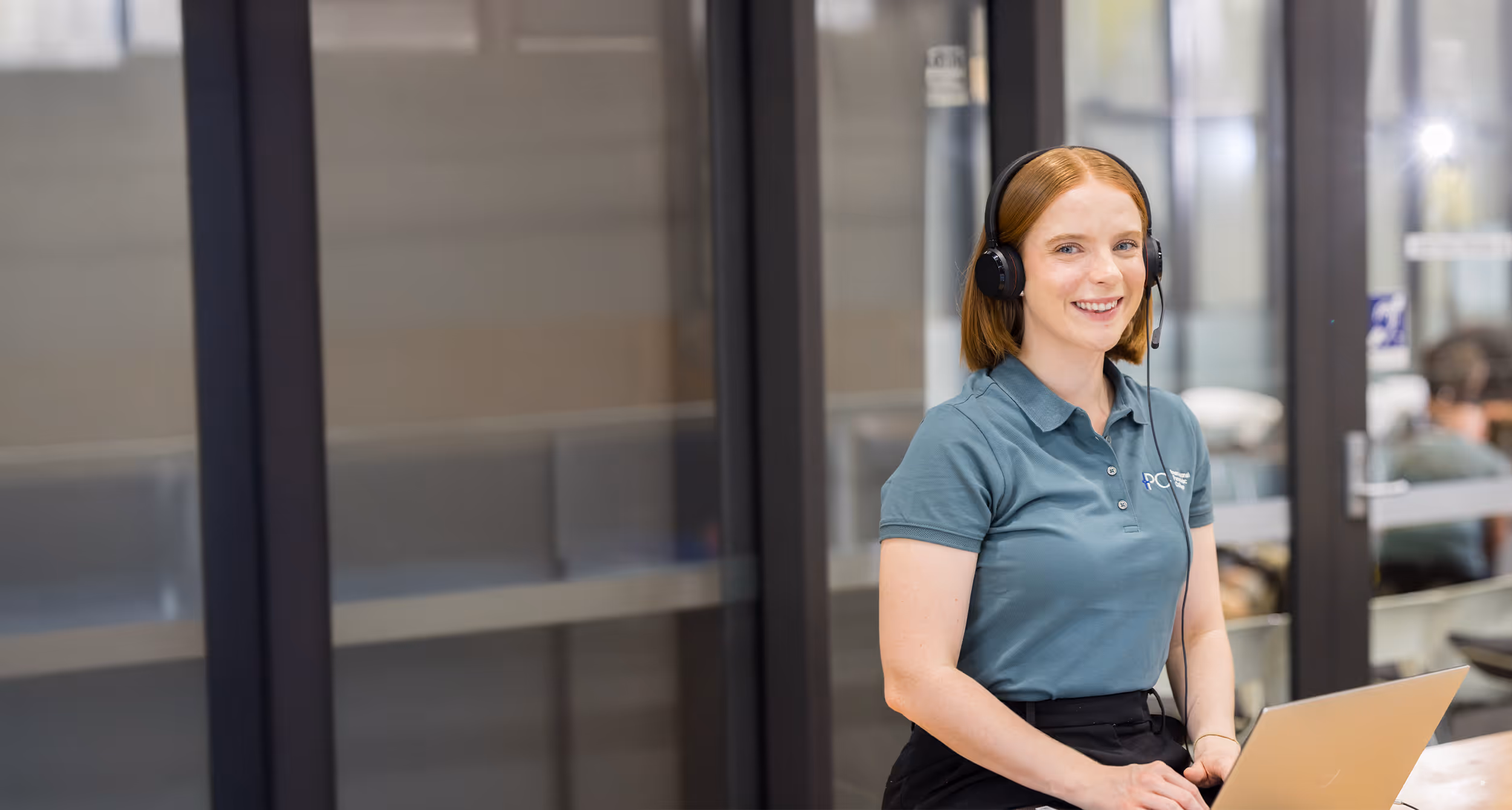 Smiling woman with red hair wearing a headset and a blue polo shirt sitting with a laptop in a bright office.