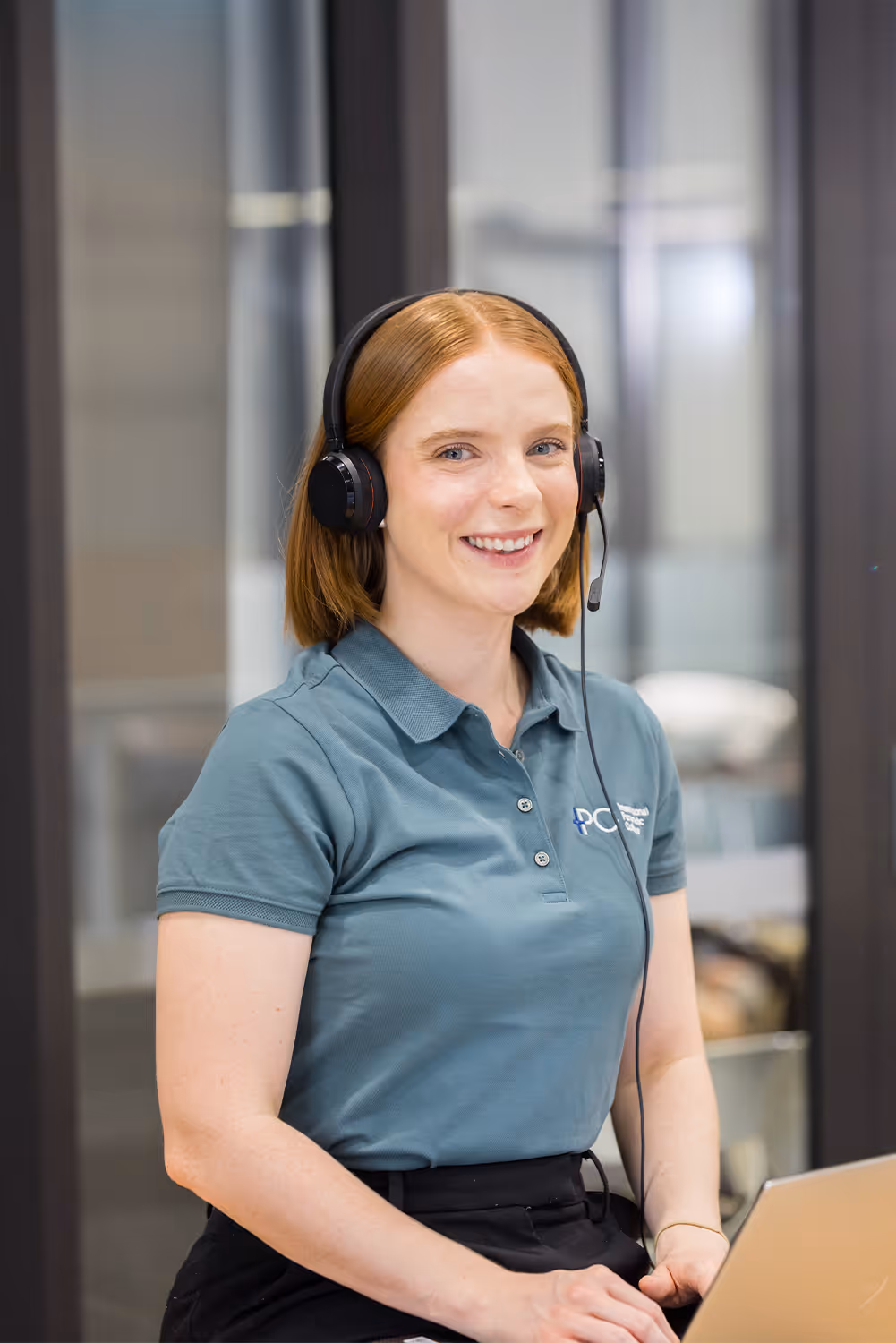 Smiling woman wearing a headset and teal polo shirt sitting in front of a laptop in an office.