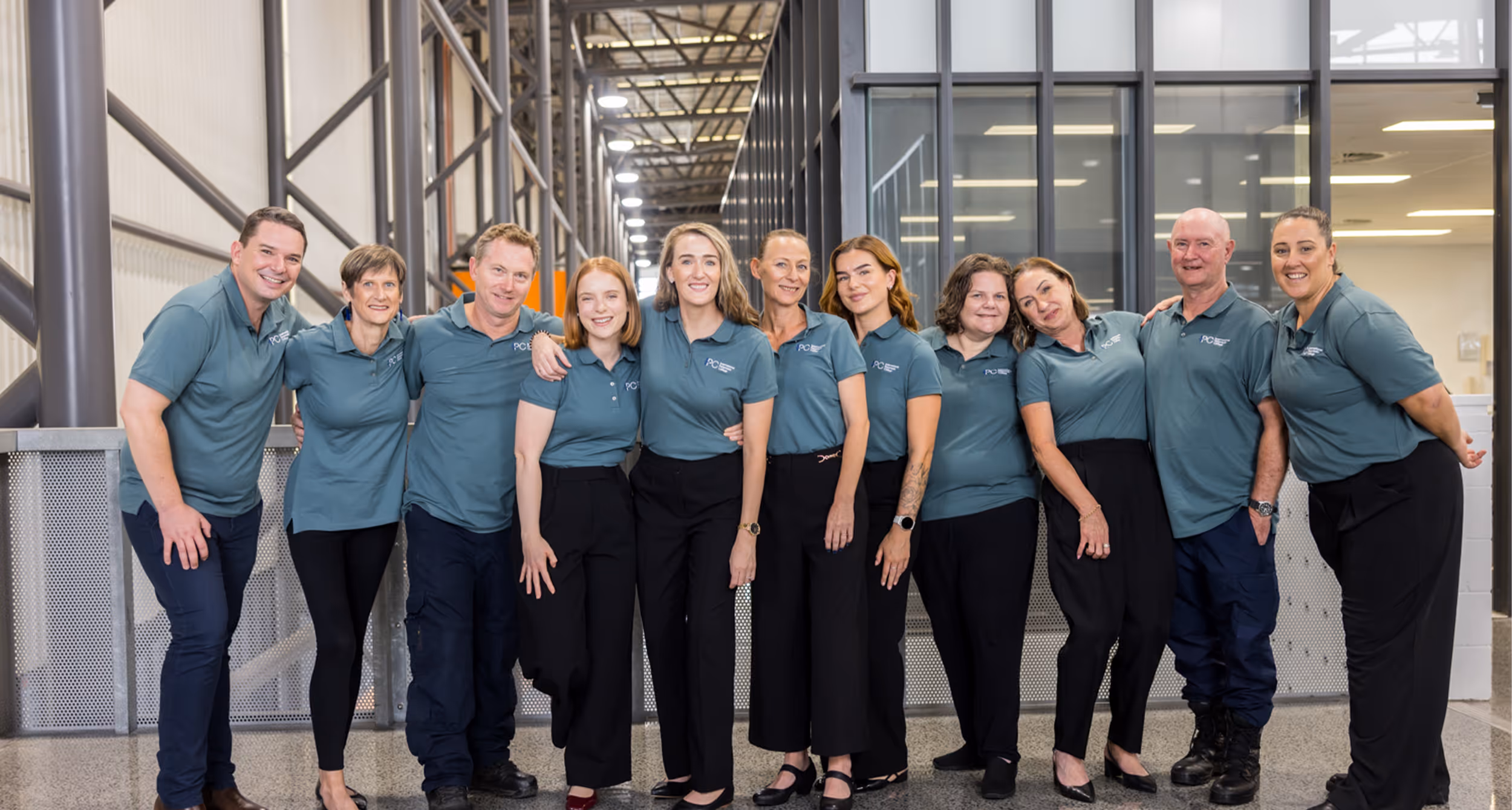 Group of eleven people wearing matching blue polo shirts and black pants standing indoors in a modern industrial office space.