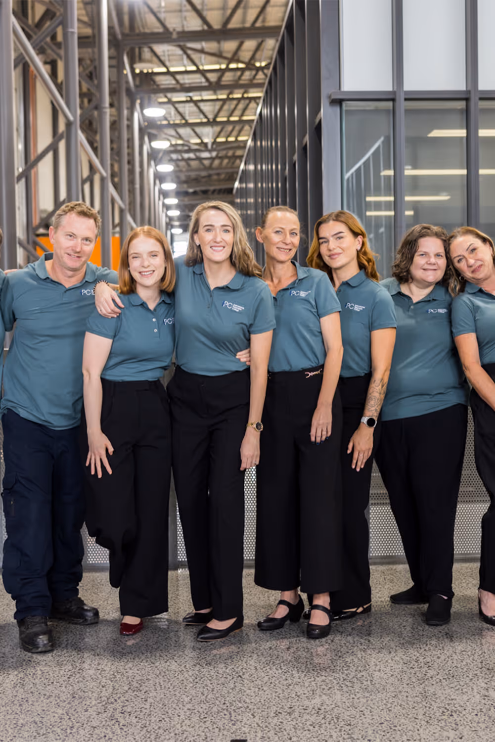 Seven colleagues standing arm in arm indoors, wearing matching teal polo shirts and black pants.