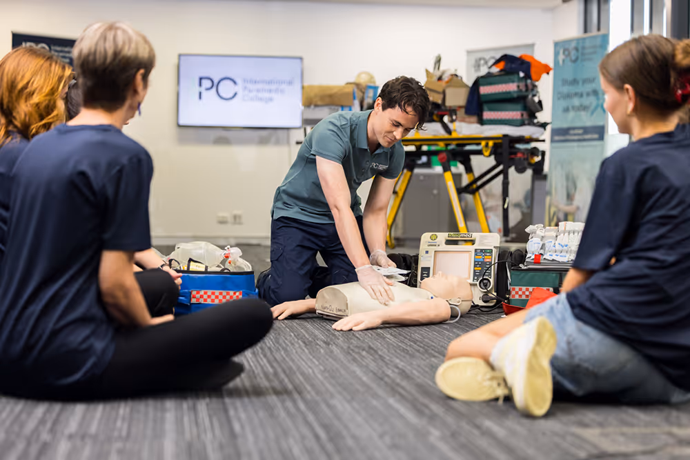 Instructor demonstrating CPR on a training mannequin while several students sit around observing during a first aid class at a training facility.