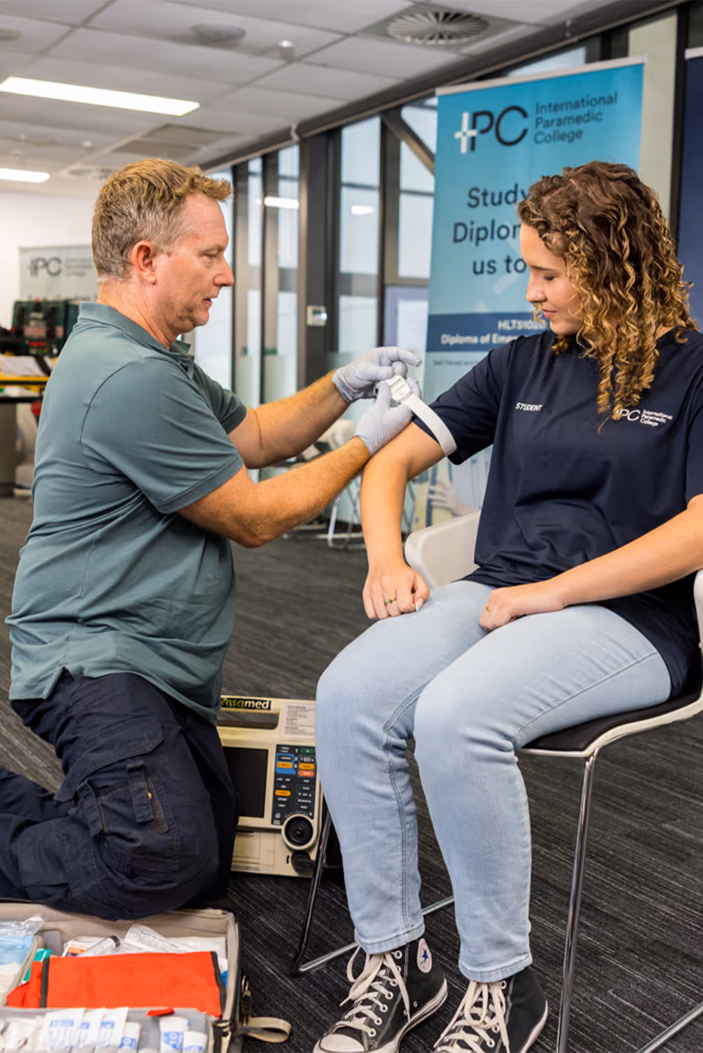 Instructor applying a tourniquet to a student’s arm during a first aid training session in a classroom with medical equipment and signage in the background.