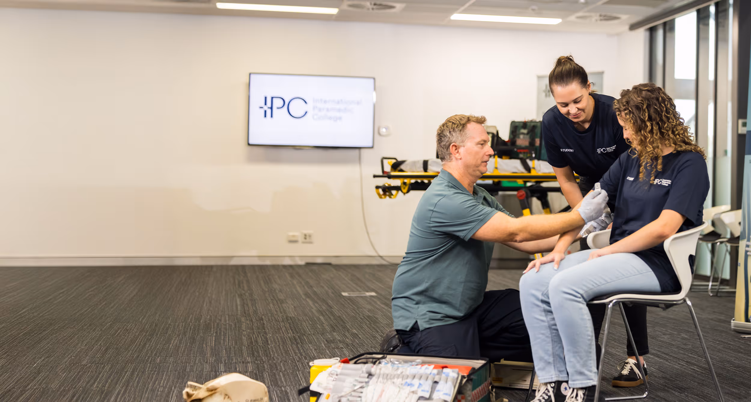 A trainer kneels beside a seated woman, demonstrating how to bandage her forearm, while another student observes and assists in a classroom first aid training setting.