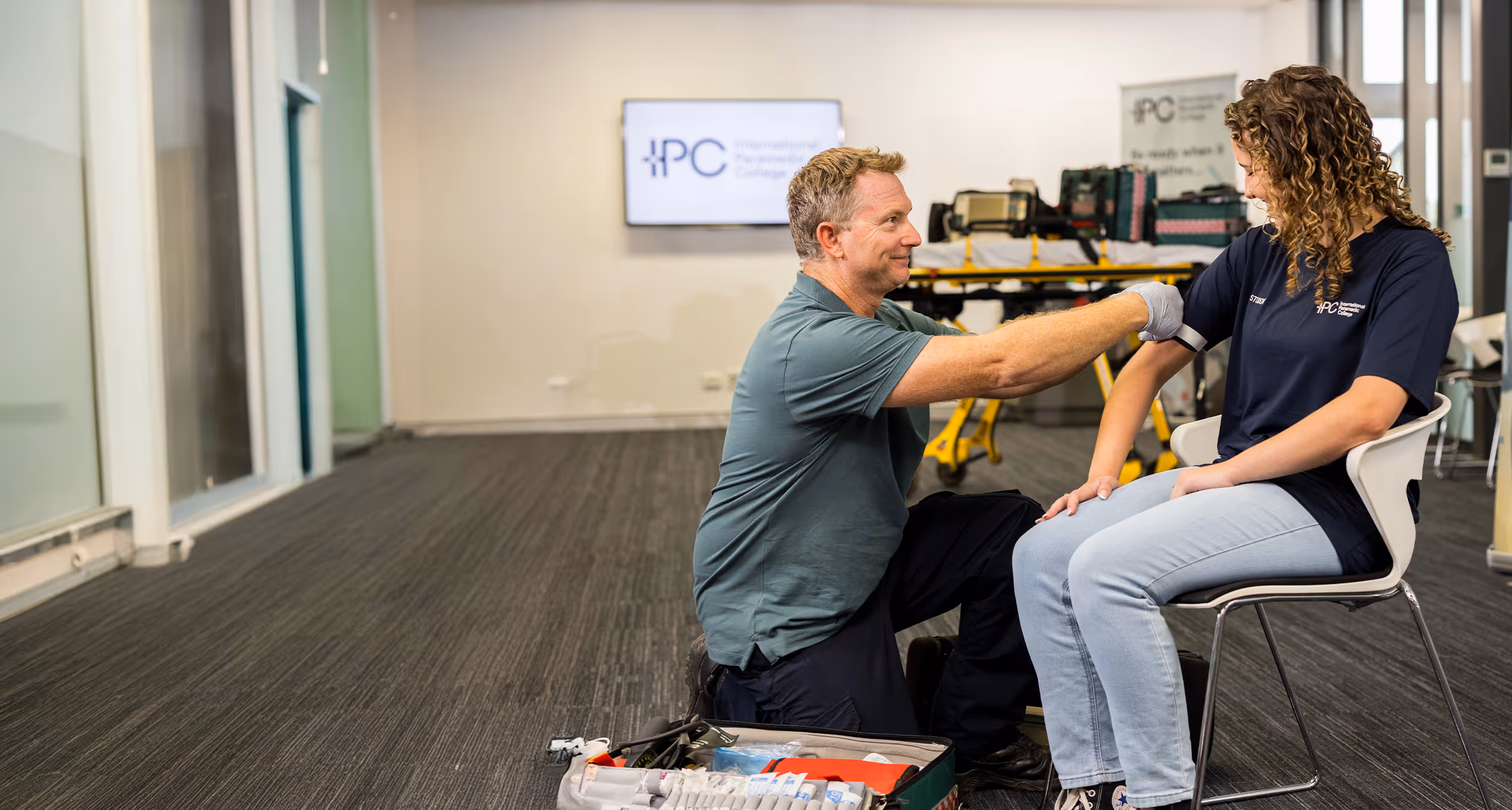 A first aid instructor kneels beside a seated student, demonstrating how to apply a tourniquet to her upper arm during a Stop the Bleed training course.