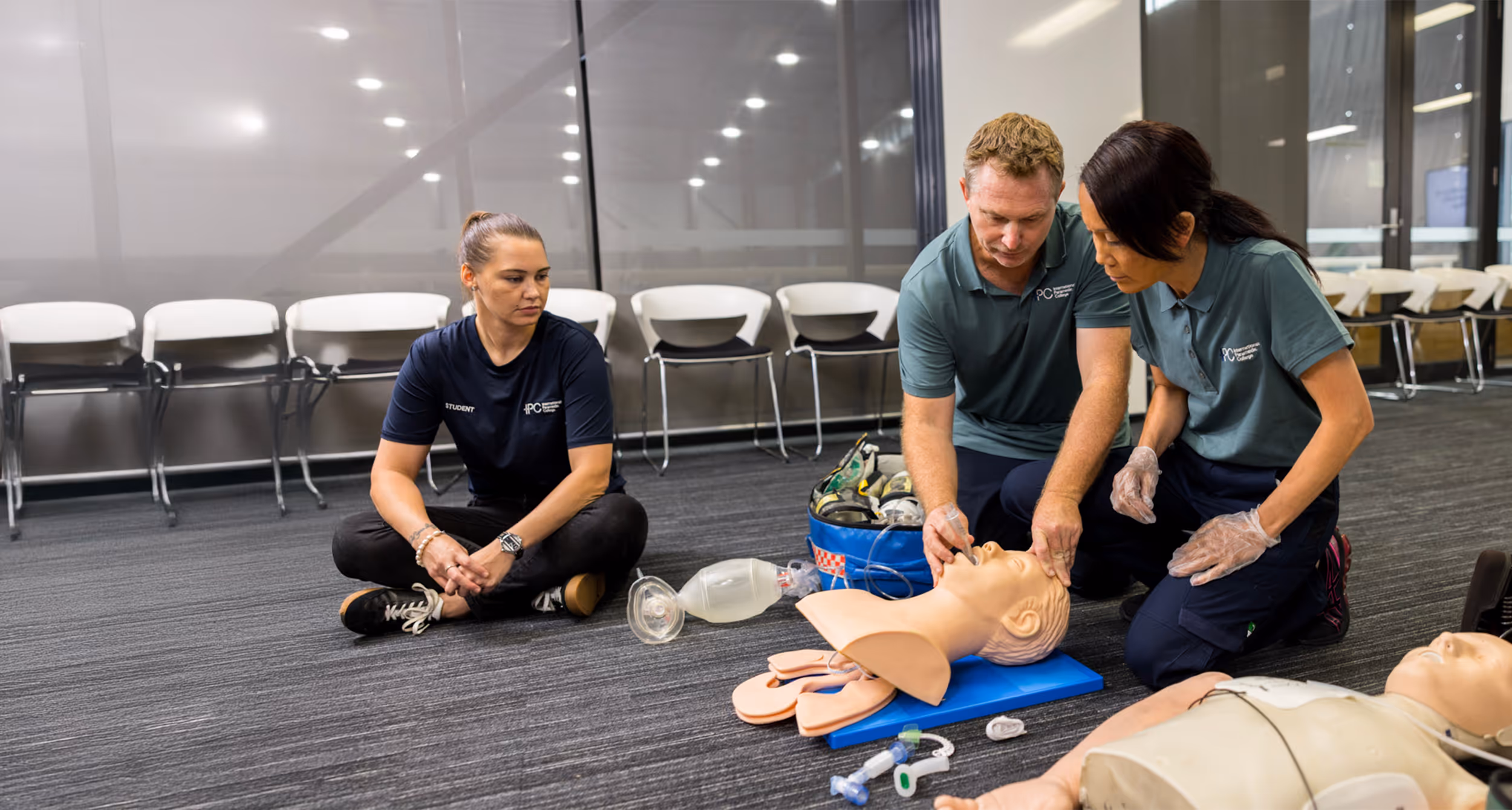 Two instructors demonstrate airway management on a training mannequin while a student watches during an advanced first aid course in a classroom setting.
