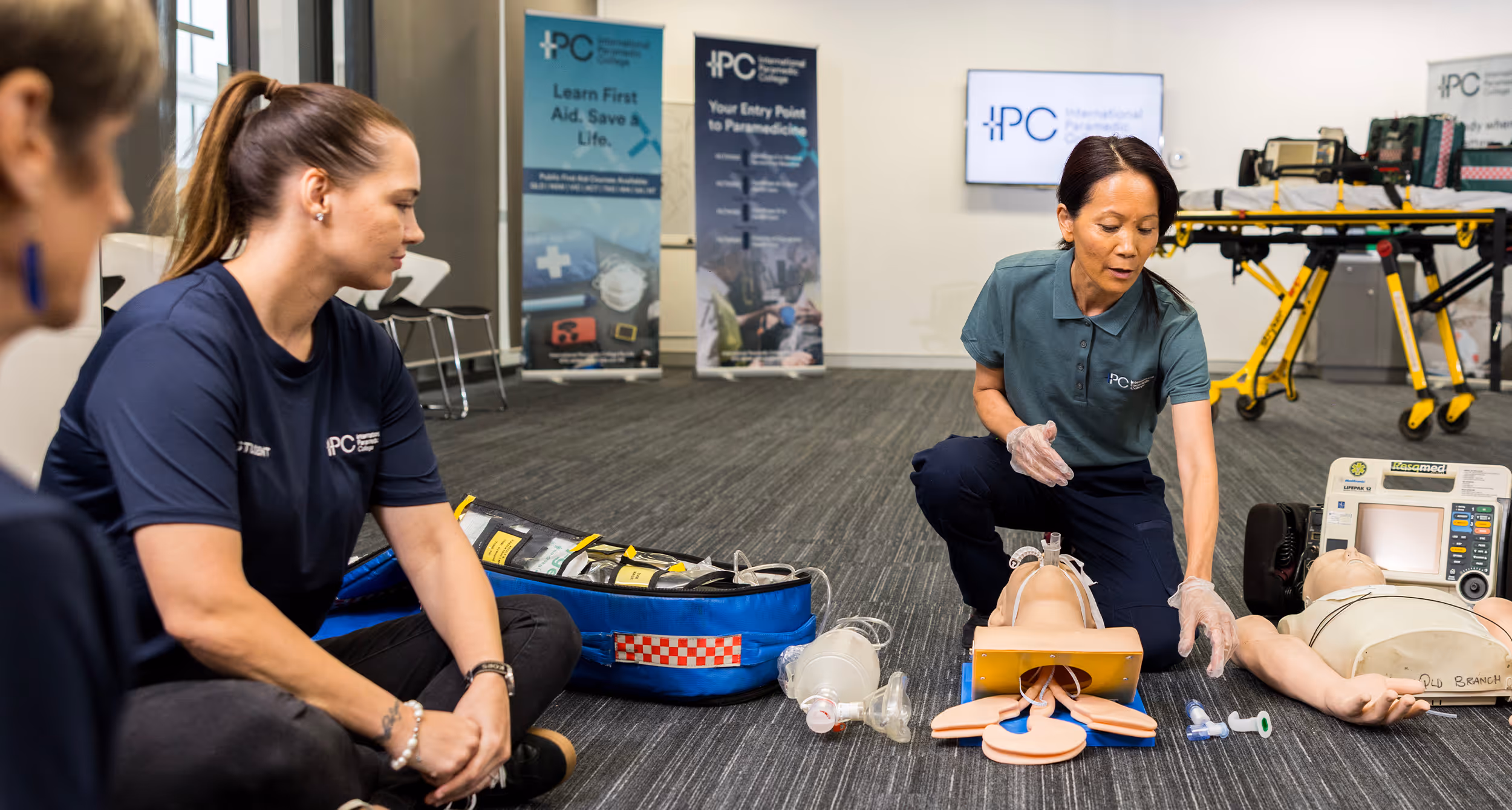 Instructor kneeling on the floor demonstrating airway management on a medical training mannequin, with resuscitation equipment arranged nearby in a classroom setting.