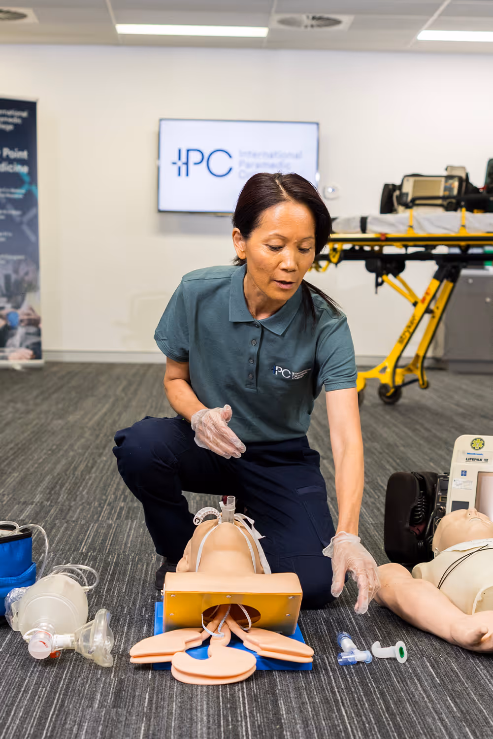 Instructor kneeling on the floor demonstrating airway management on a medical training mannequin, with resuscitation equipment arranged nearby in a classroom setting.
