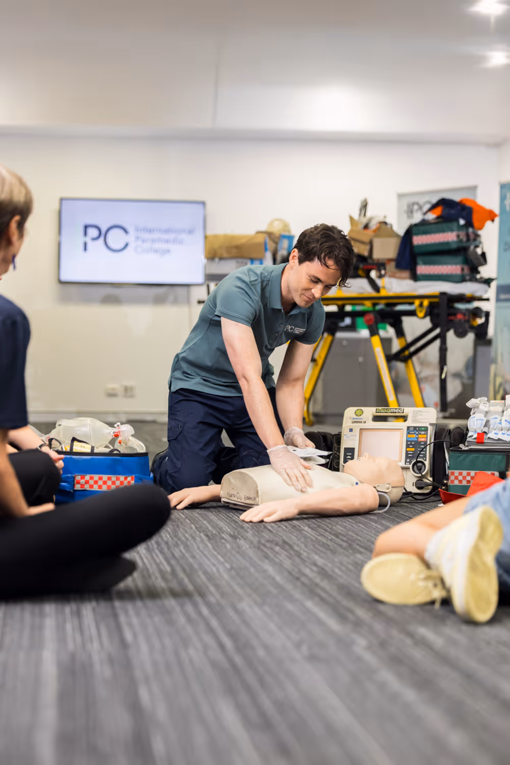 Instructor performing CPR chest compressions on a training mannequin while students observe during a paramedic training session.