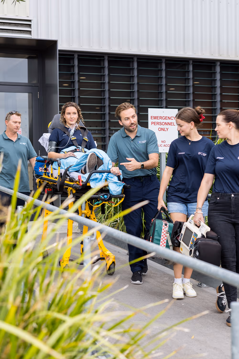 Instructor walking with paramedic students as they transport a patient on a stretcher and carry medical equipment during an outdoor training exercise.