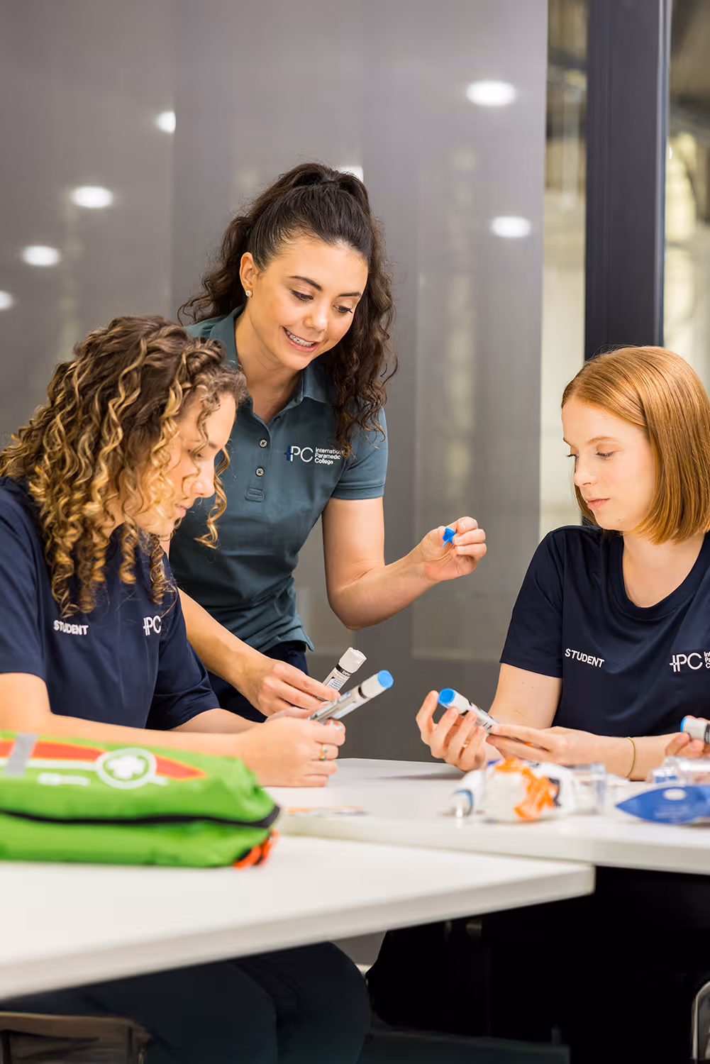 Instructor supervising two students as they practice using medical devices at a classroom table during paramedic training.