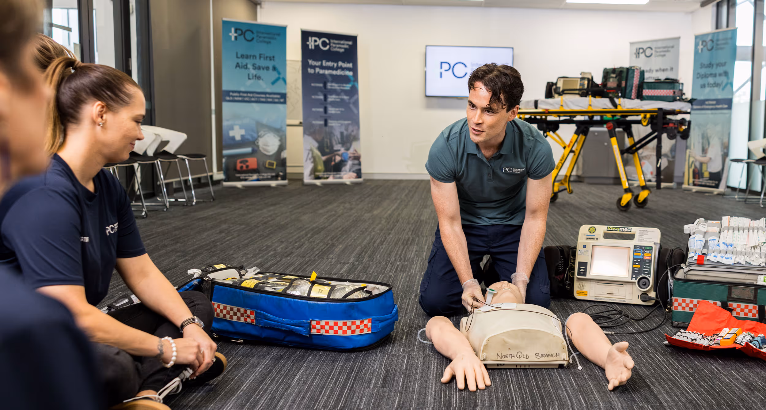 Instructor performing CPR compressions on a training mannequin with a defibrillator and medical kit nearby in a classroom setting.