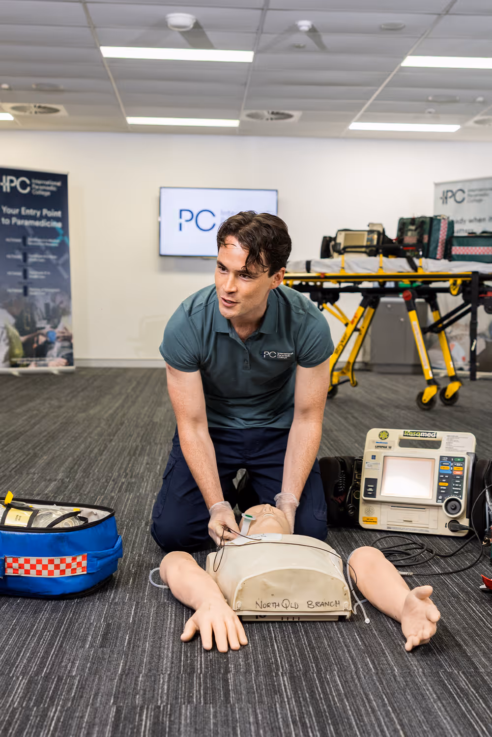 Instructor performing CPR compressions on a training mannequin with a defibrillator and medical kit nearby in a classroom setting.