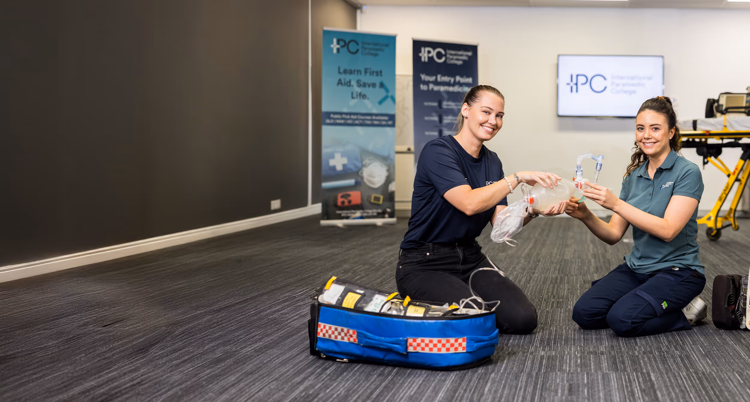 Instructor and student kneeling on the floor practicing ventilation techniques with a bag-valve mask during a paramedic training session.