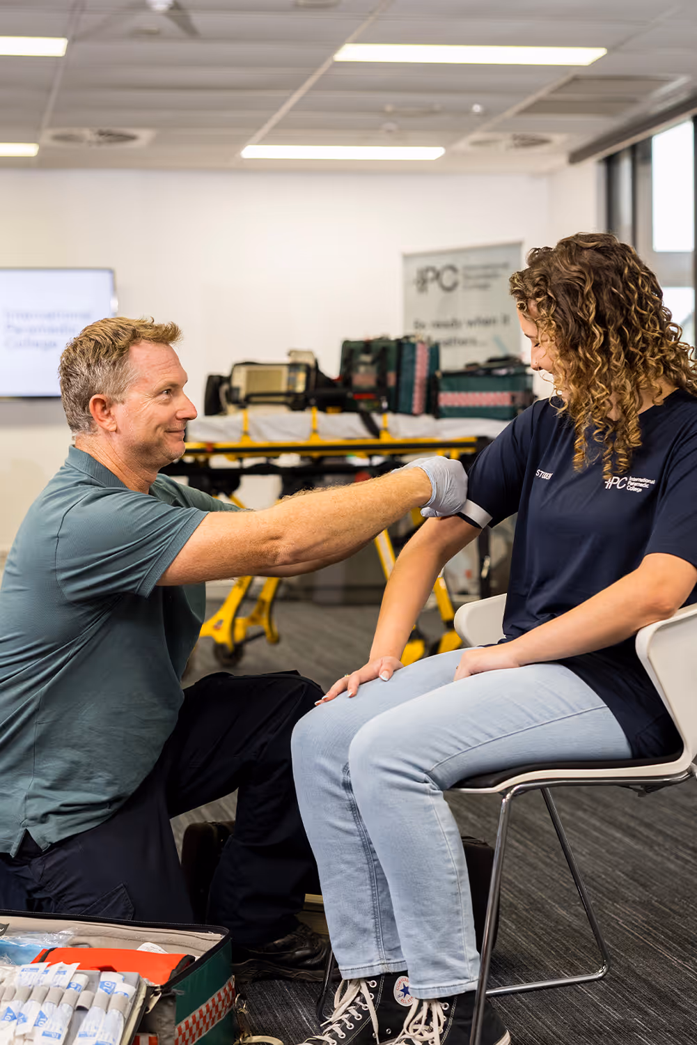 Instructor applying a tourniquet to a student’s arm during a first aid training session, with medical supplies visible nearby.