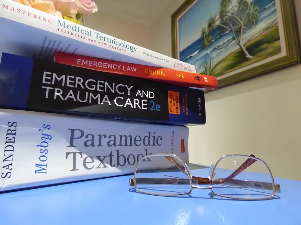 Stack of paramedic and emergency care textbooks with reading glasses, featuring the International Paramedic College logo
