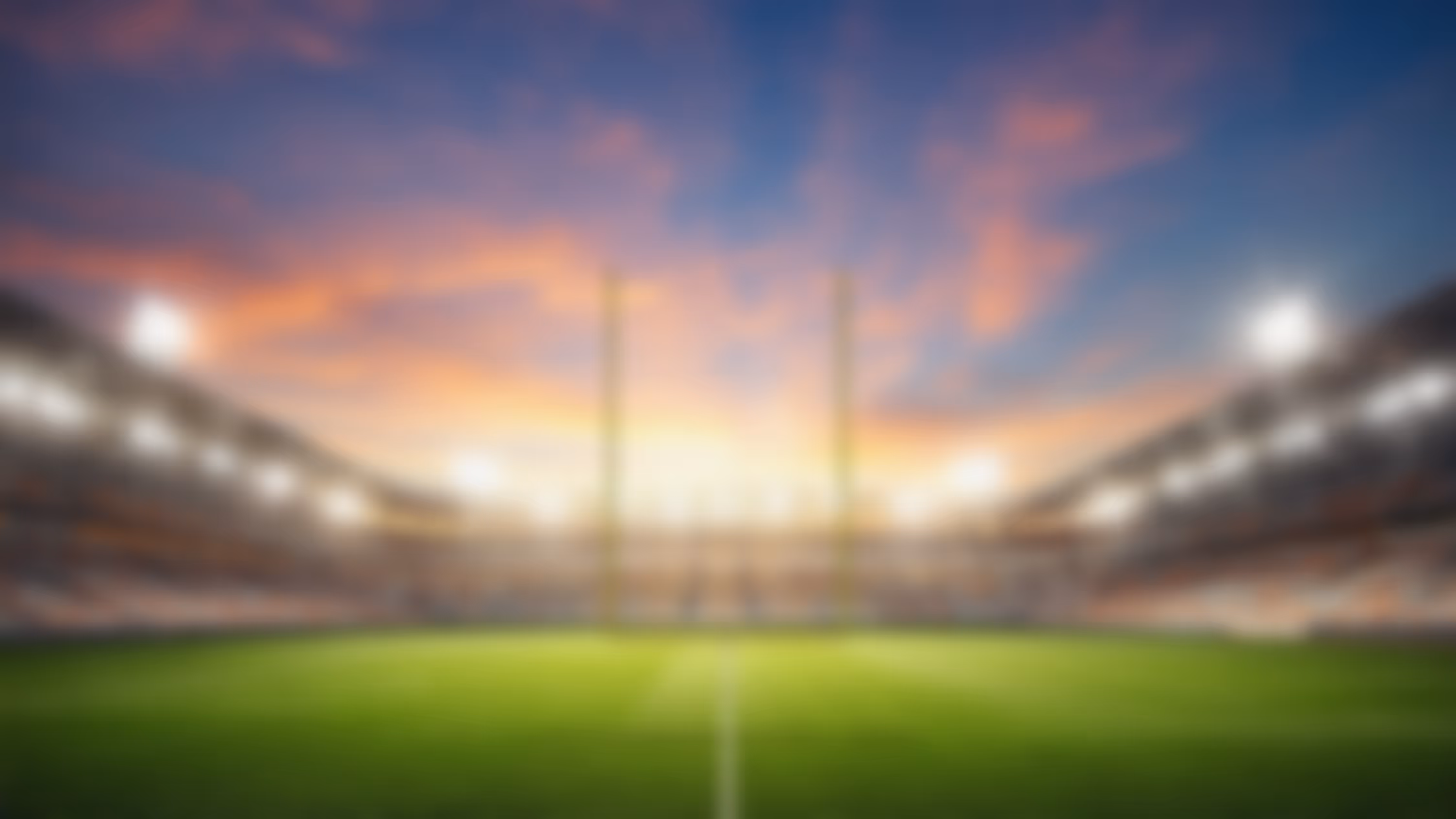 Blurred view of a stadium with goalposts, bright lights, and a sunset sky.
