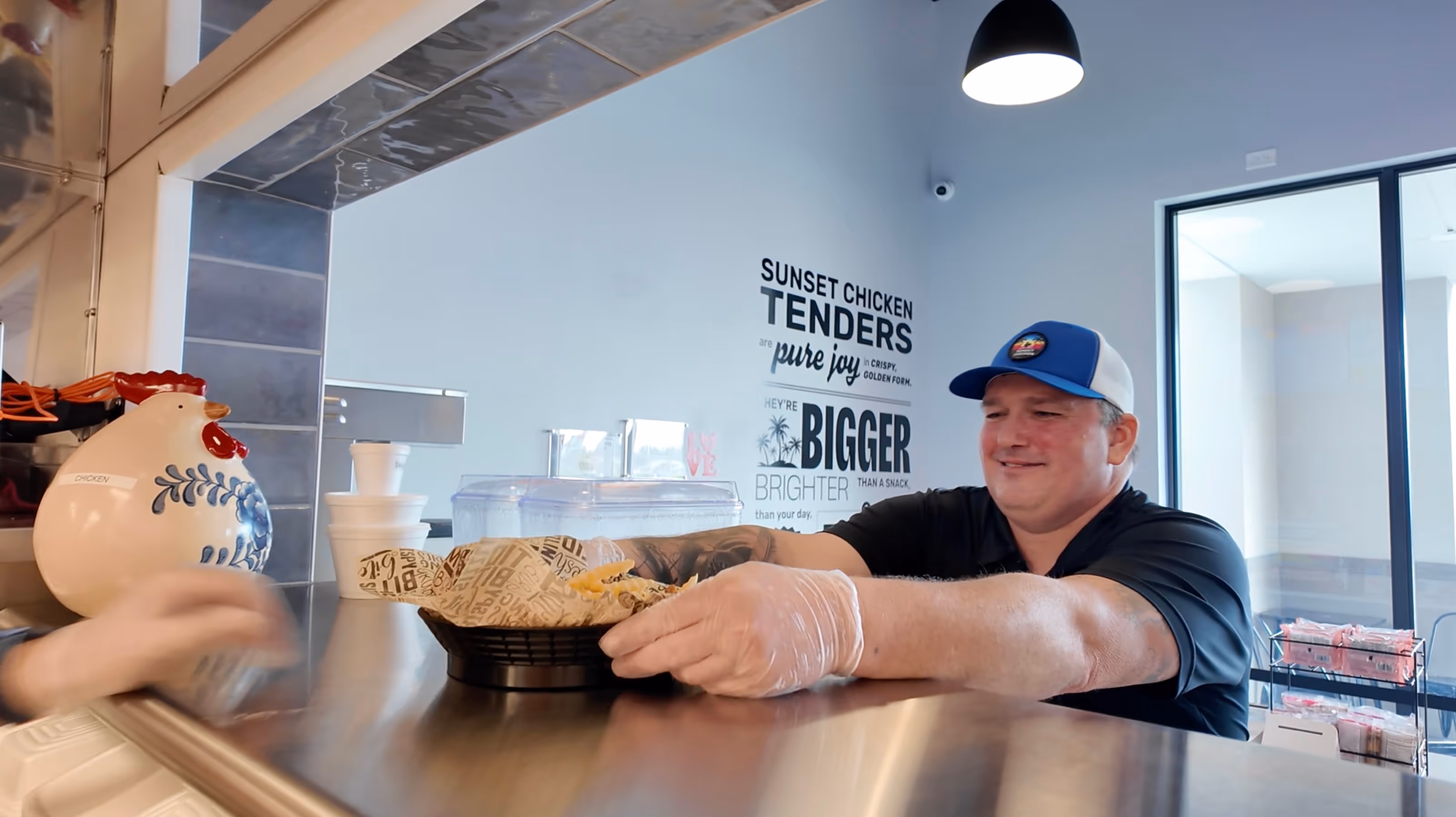 Man wearing a blue and white cap handing a basket of fries across a counter in a restaurant with a chicken-themed decor.