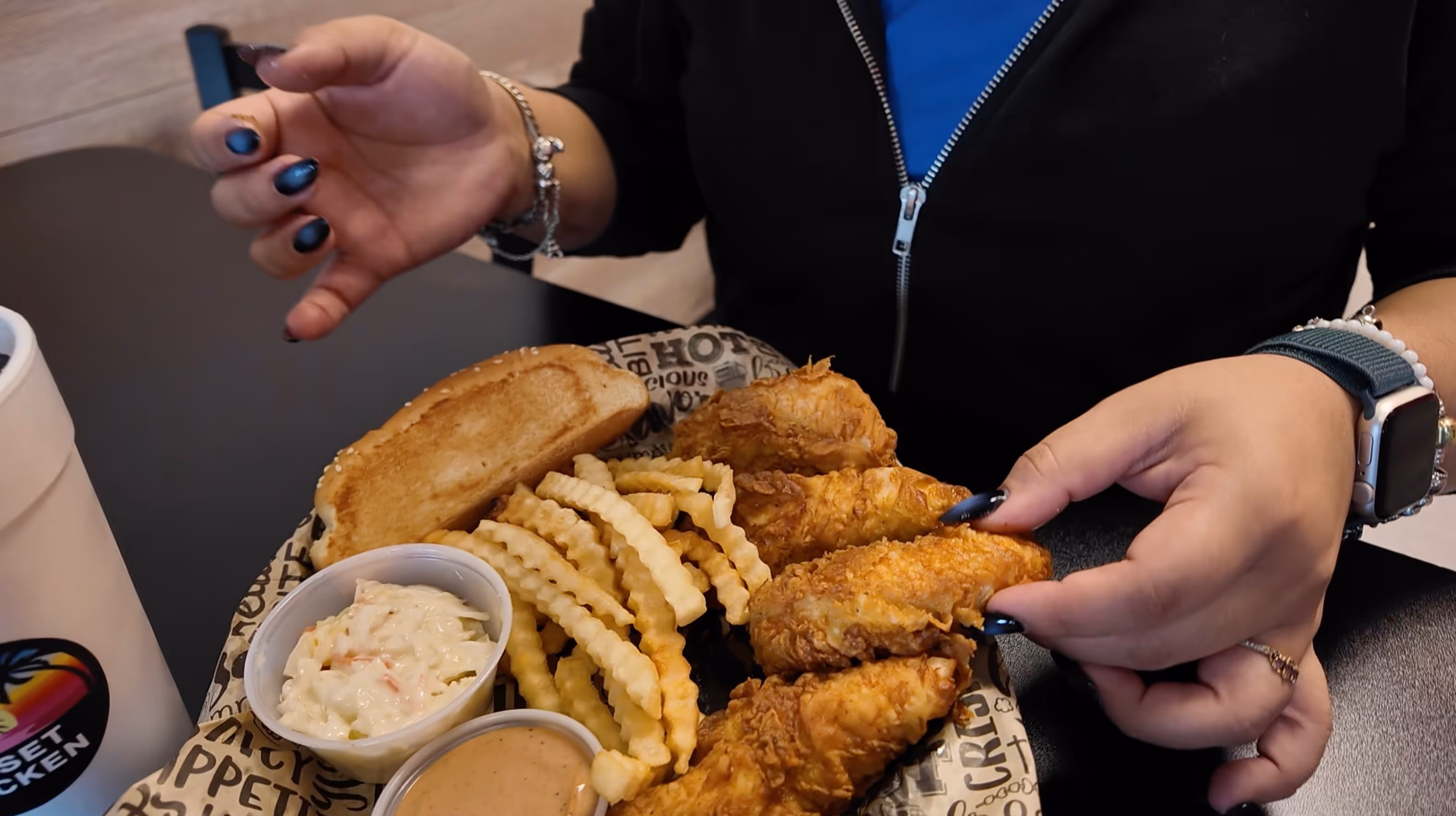 Person holding a fried chicken tender from a plate with crinkle-cut fries, toasted bun, coleslaw, and dipping sauce.
