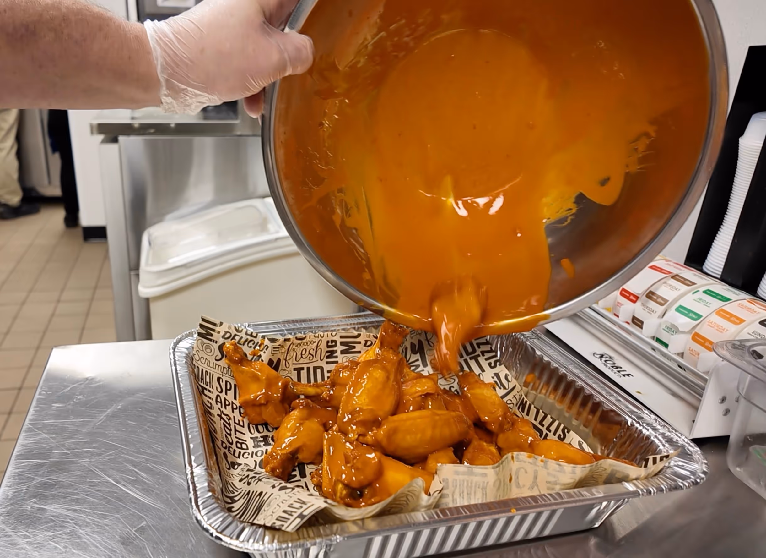 Gloved hand pouring orange sauce over cooked chicken wings in a foil tray lined with printed paper.