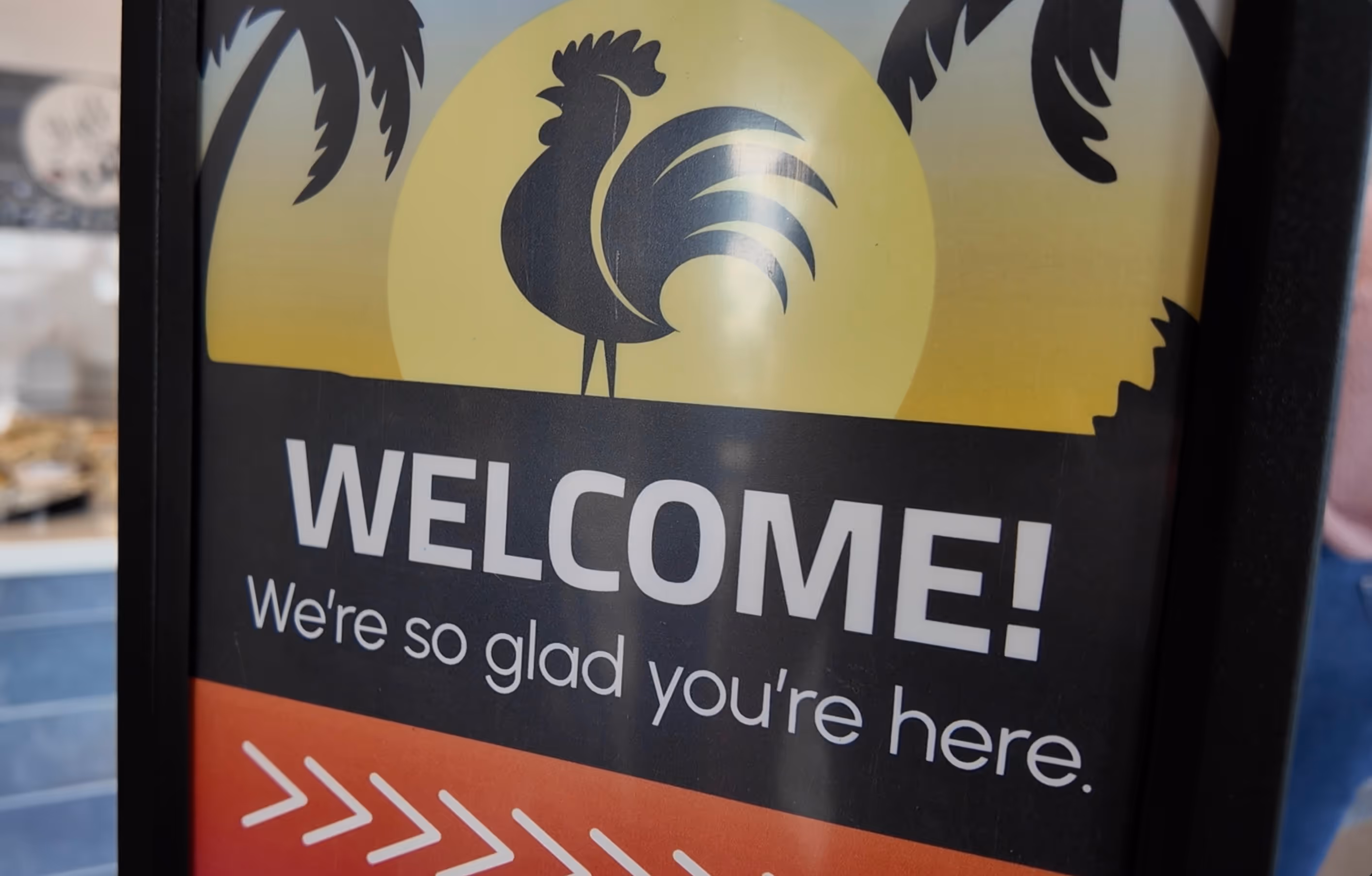 Welcome sign with a black rooster silhouette in front of a yellow sun and palm leaves, text reads 'WELCOME! We're so glad you're here.'