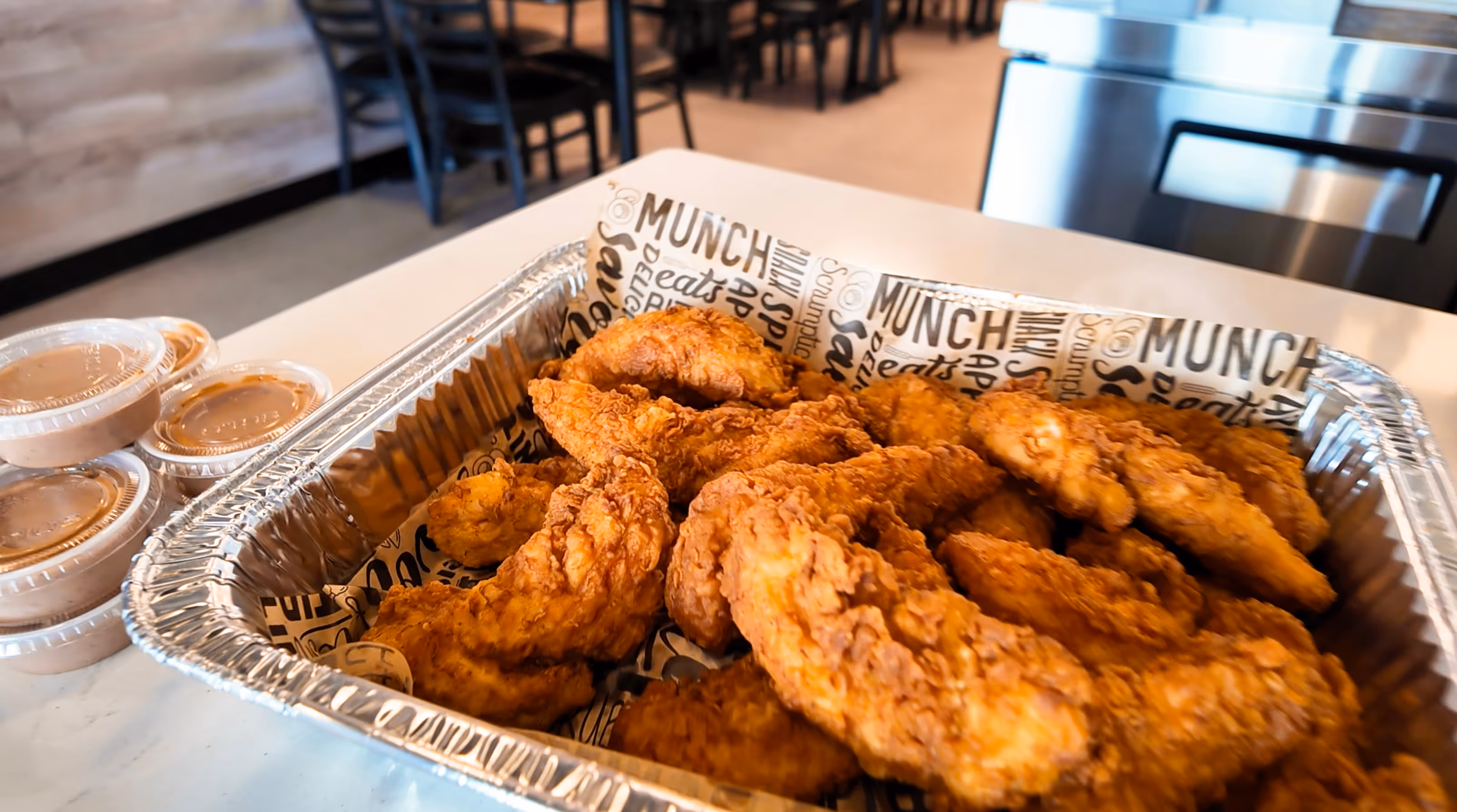 Aluminum tray filled with crispy fried chicken strips on a table with several small containers of dipping sauce beside it.