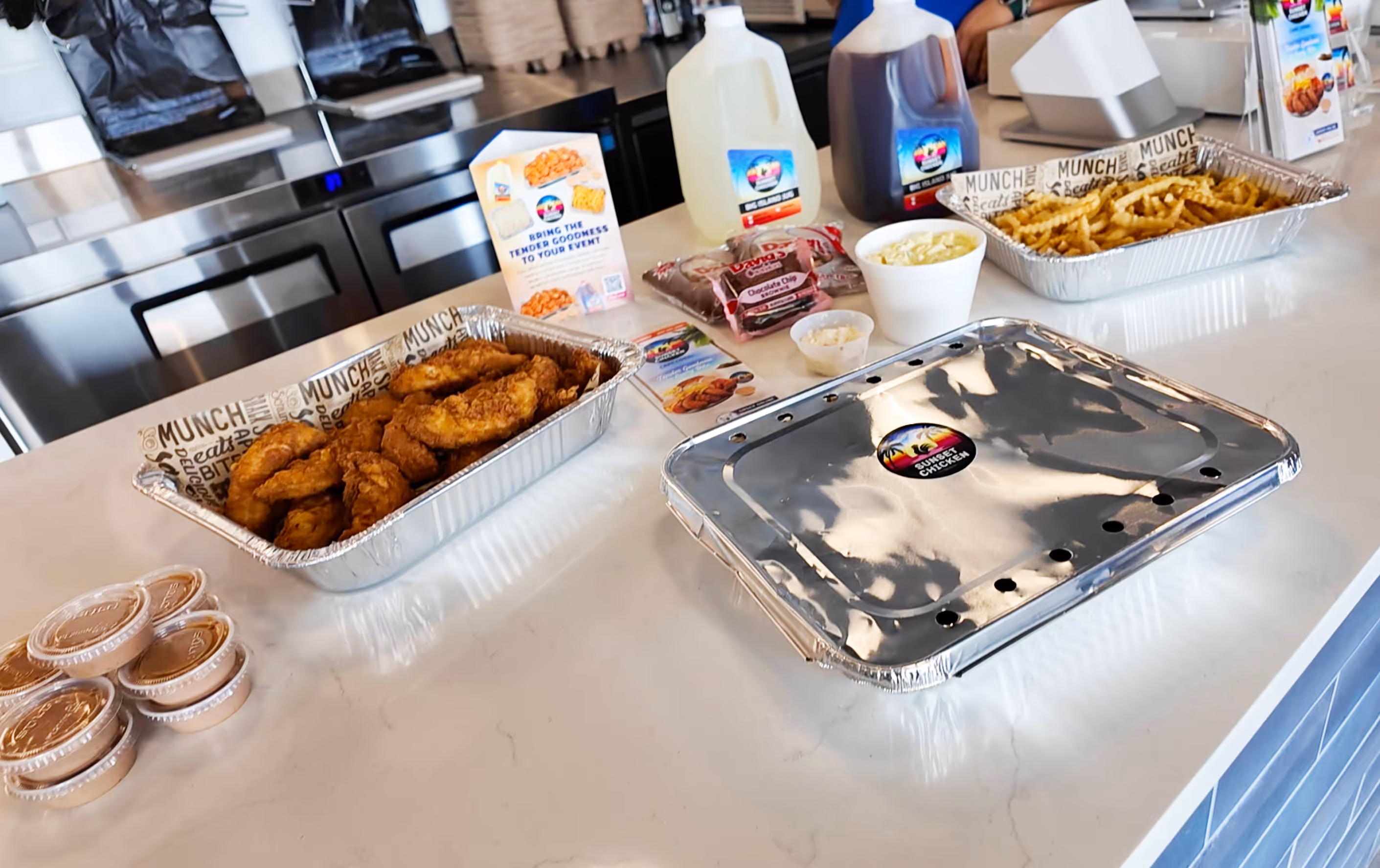 Catering spread with fried chicken tenders, crinkle-cut fries, dipping sauces, two large drink jugs, and packaged desserts on a countertop.