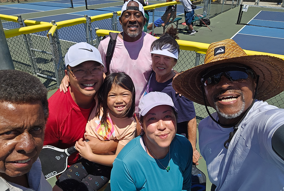 A diverse group of smiling people, including a child, posing for a selfie at an outdoor pickleball court on a sunny day.