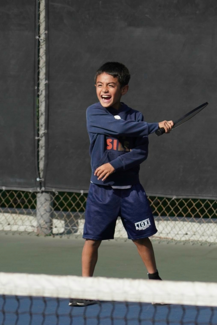 Smiling boy in navy sweatshirt and shorts holding a tennis racket on a tennis court.
