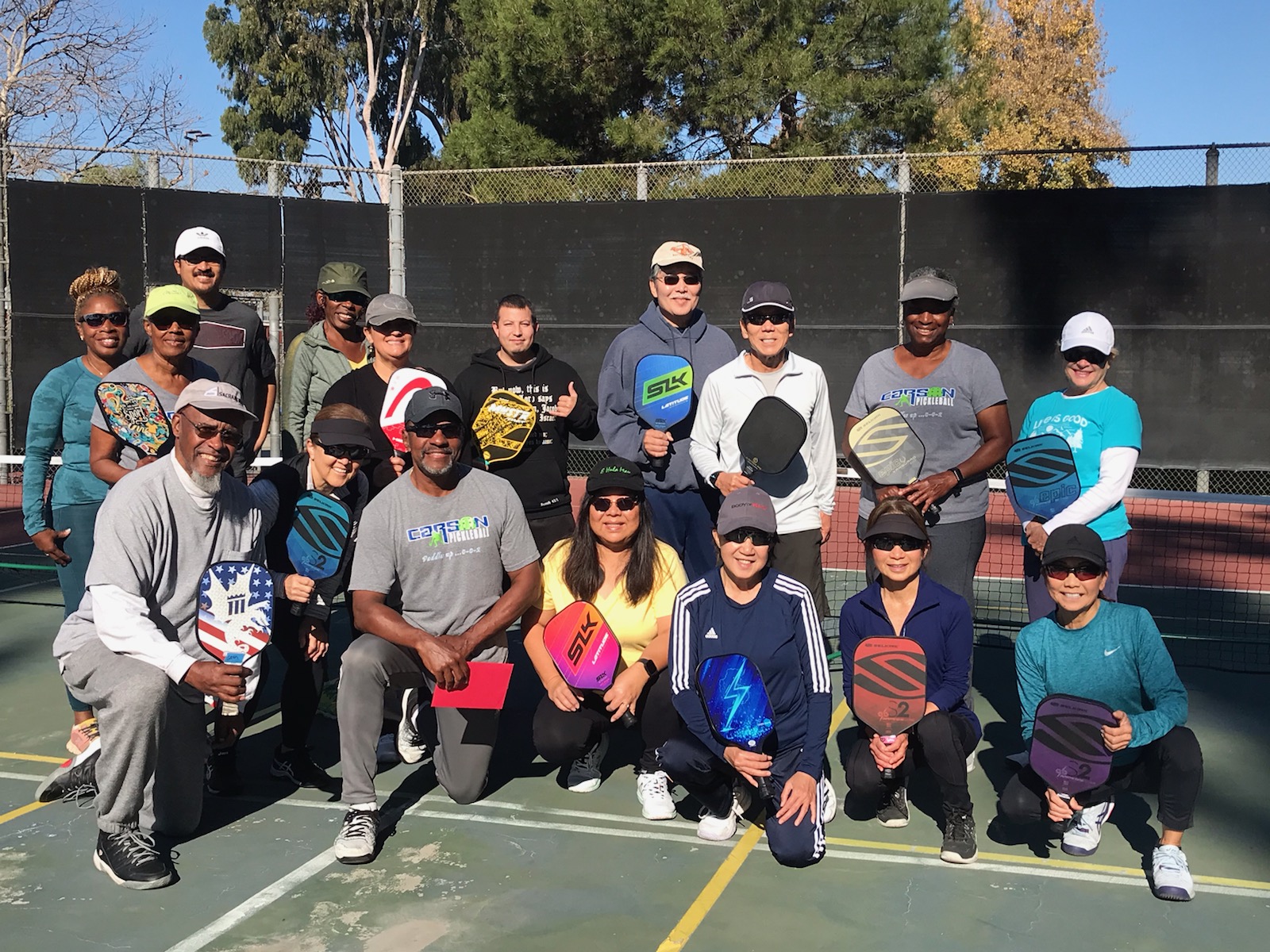 Group of 18 adult pickleball players posing on a court holding paddles, with trees and a fence in the background.
