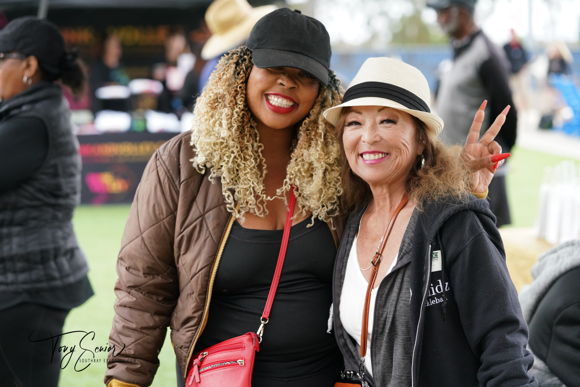 Two smiling women posing together, one with curly blonde hair wearing a black cap and brown jacket, the other with a white hat and black hoodie making a peace sign.