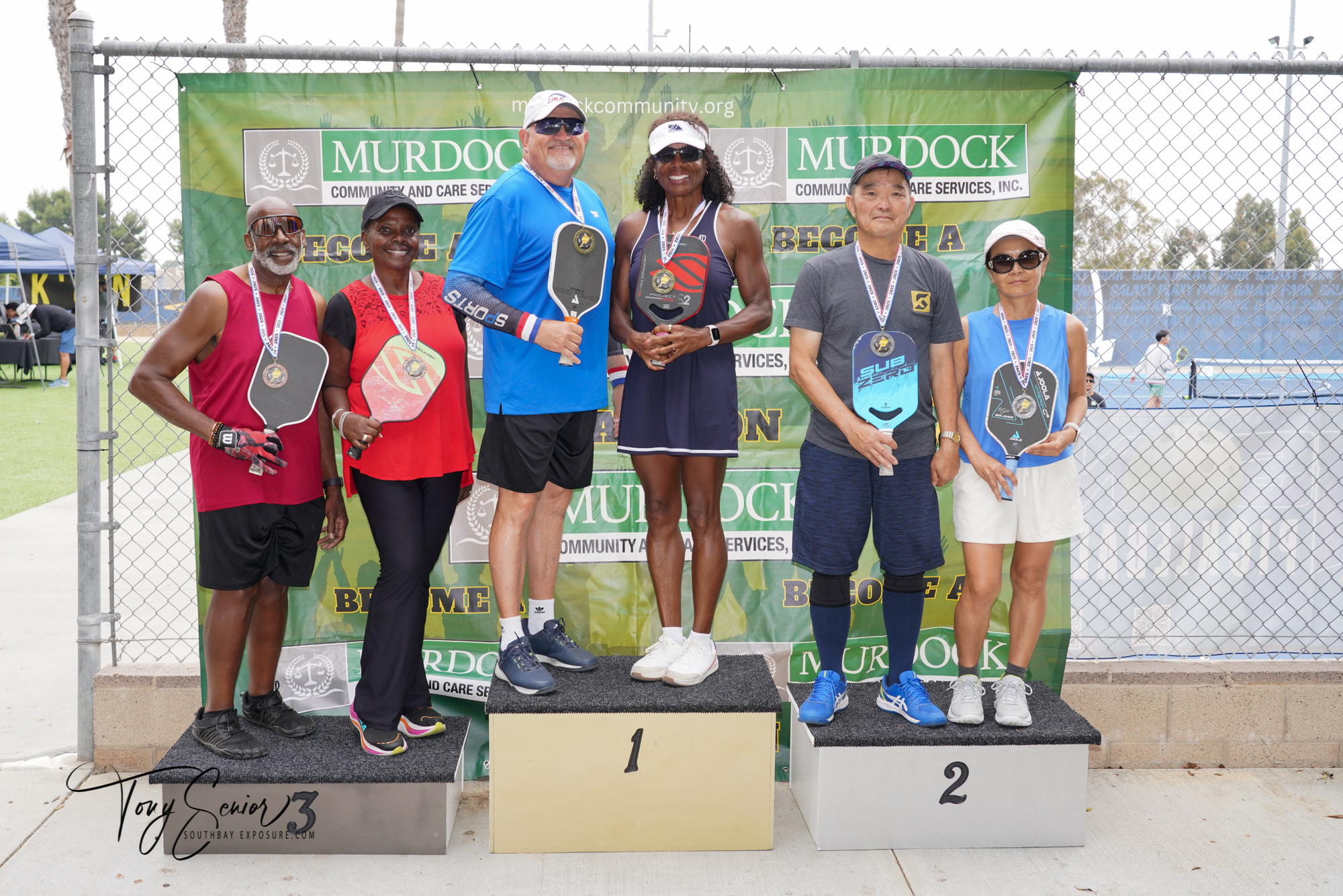 Six pickleball players wearing medals stand on winners’ podiums holding paddles in front of a Murdock Community banner.