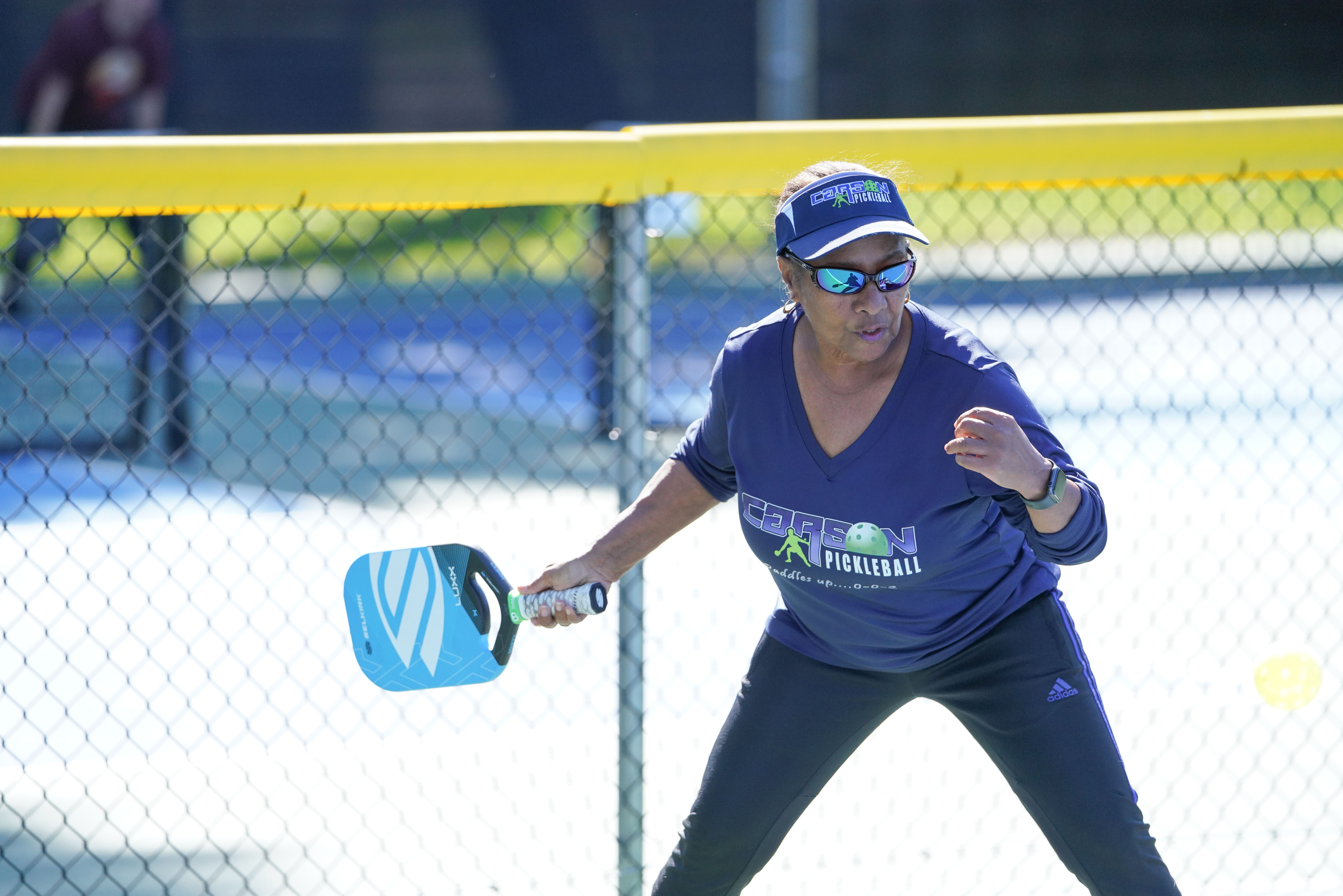 Woman wearing sunglasses and visor playing pickleball outdoors with a blue paddle.