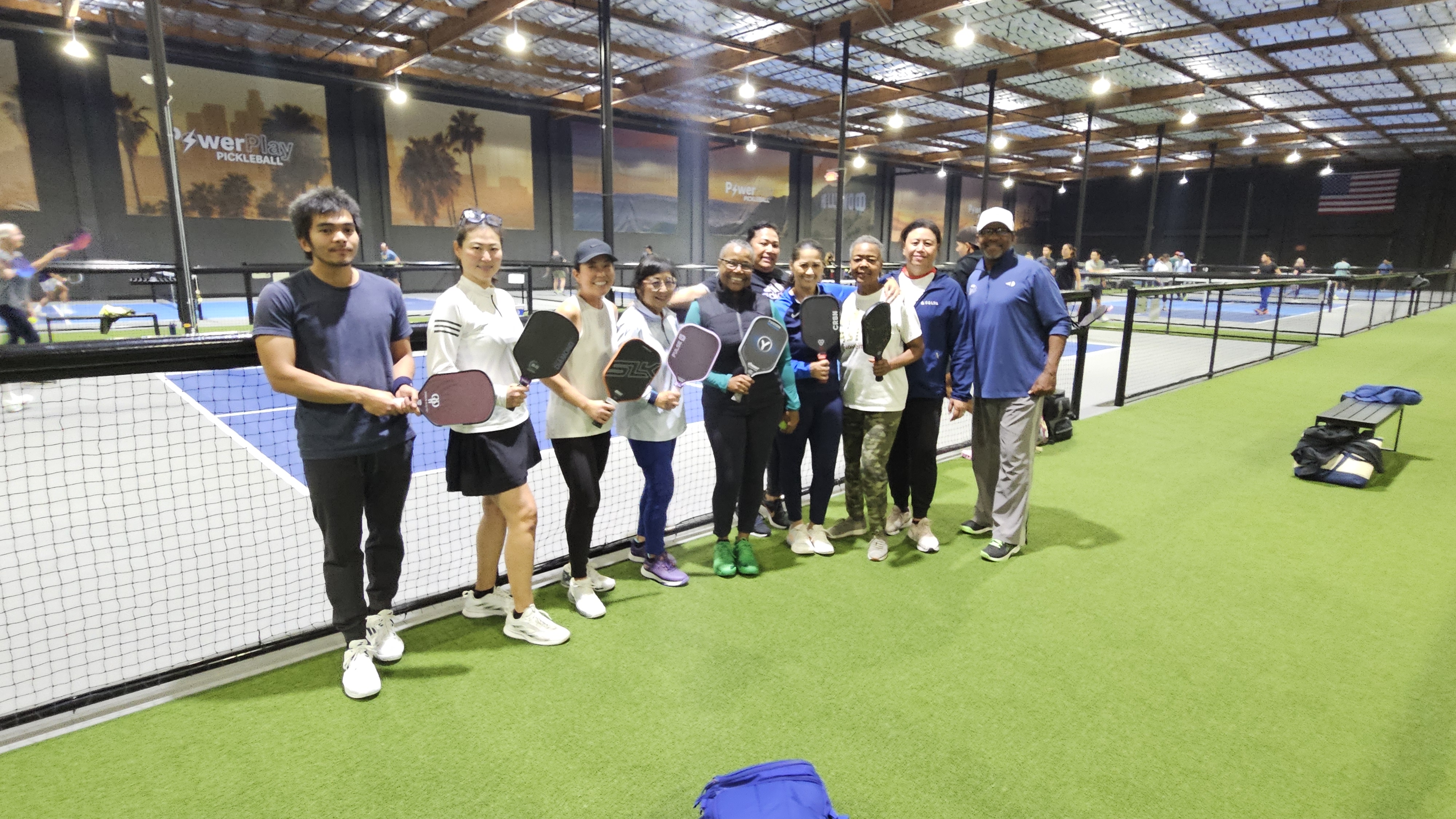 Group of ten people standing indoors on an artificial grass court holding pickleball paddles, with several pickleball courts and players in the background.