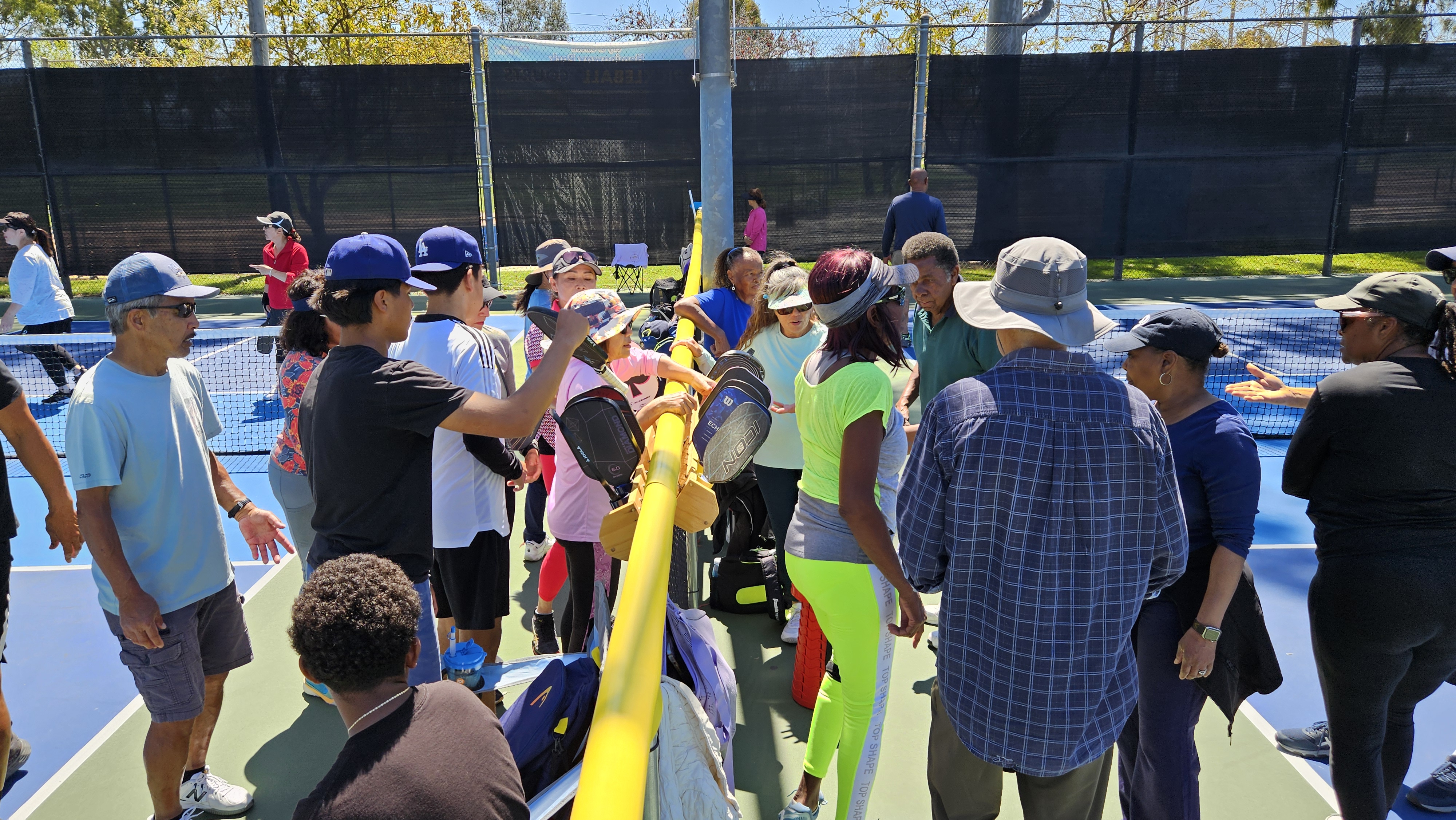 Group of people gathered on an outdoor pickleball court with paddles resting on a yellow railing.