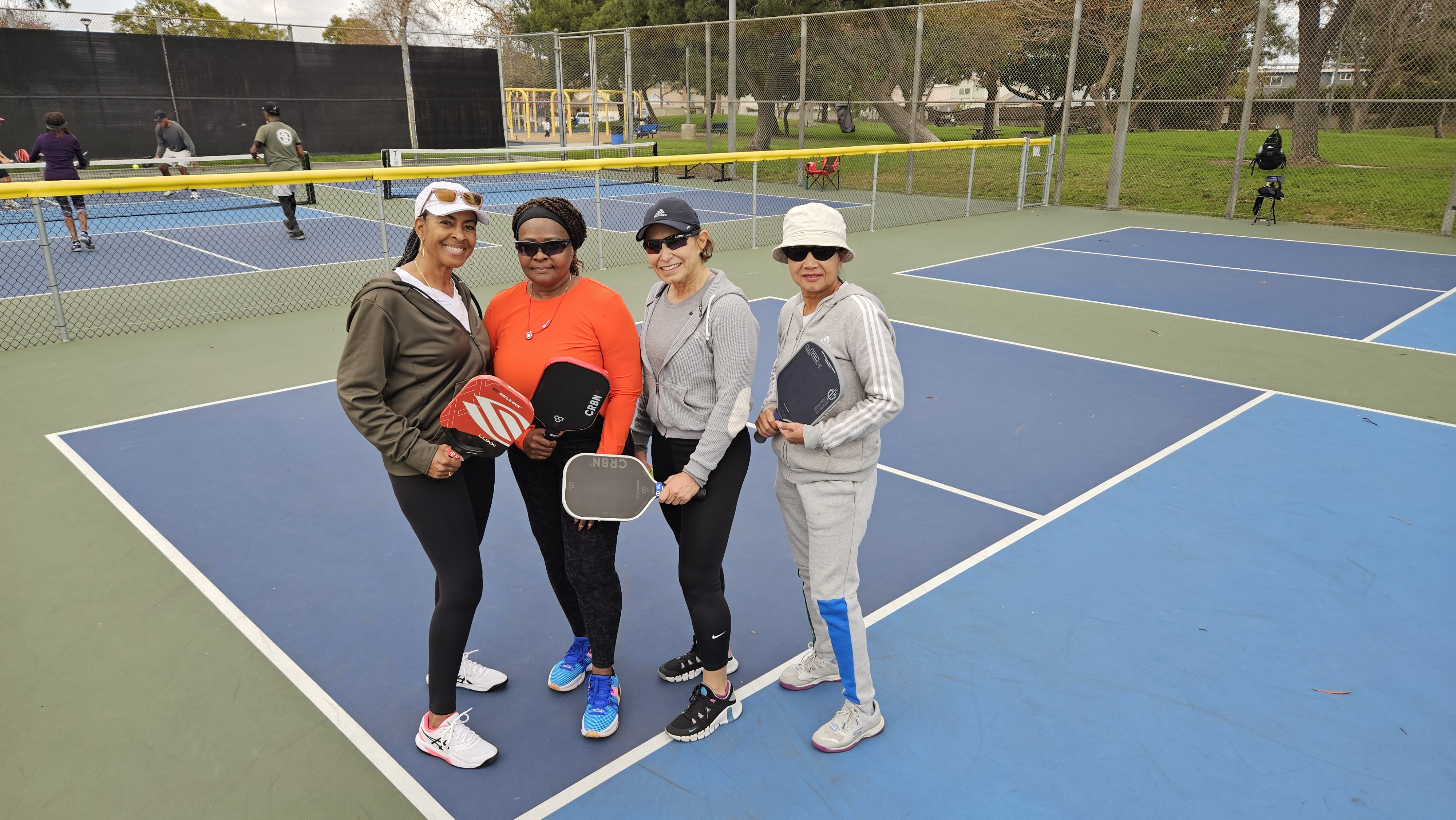 Four women posing with pickleball paddles on an outdoor pickleball court.