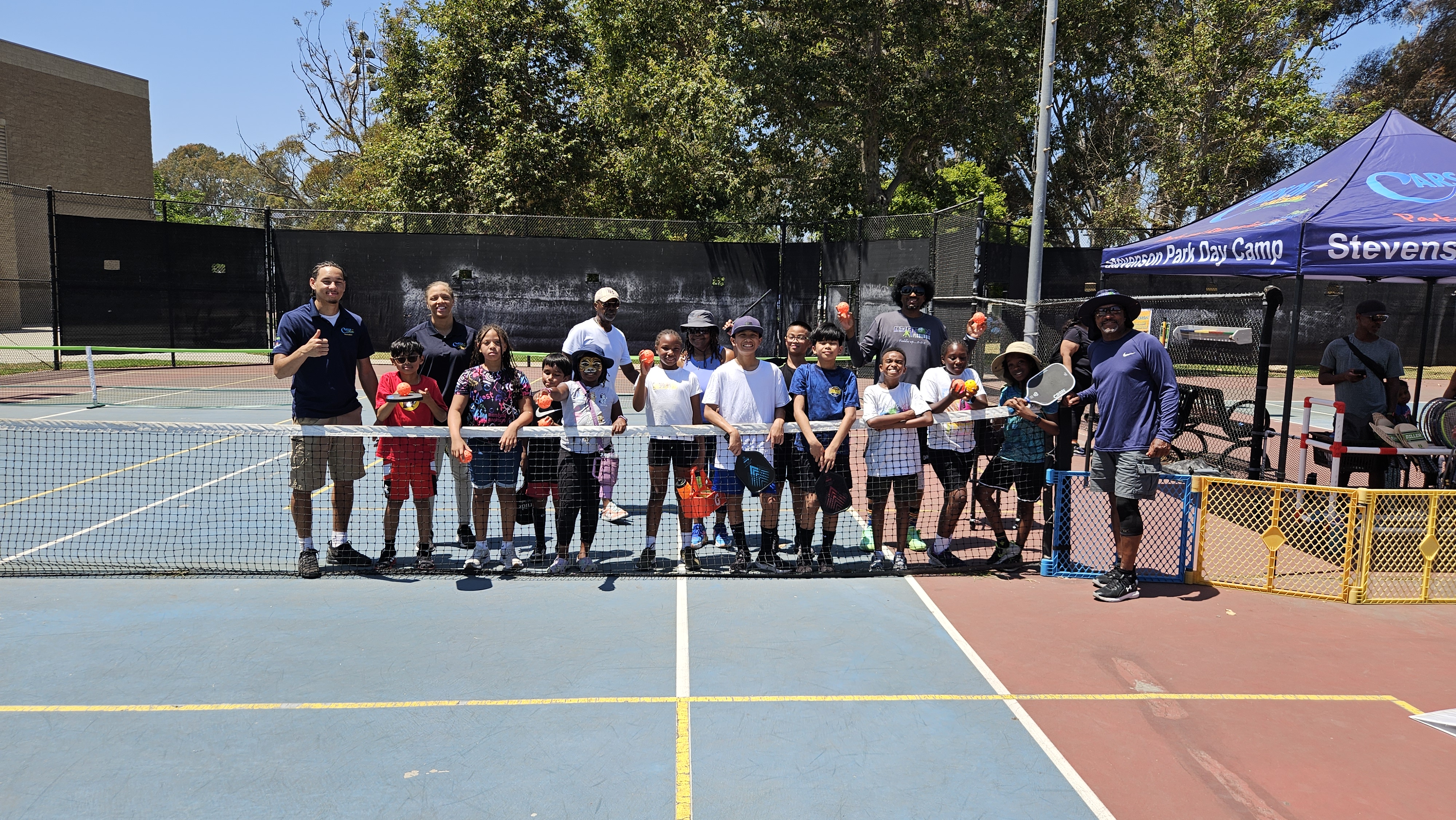 Group of children and adults standing behind a pickleball net on an outdoor court, holding paddles and balls, with a Stevenson Park Day Camp tent in the background.