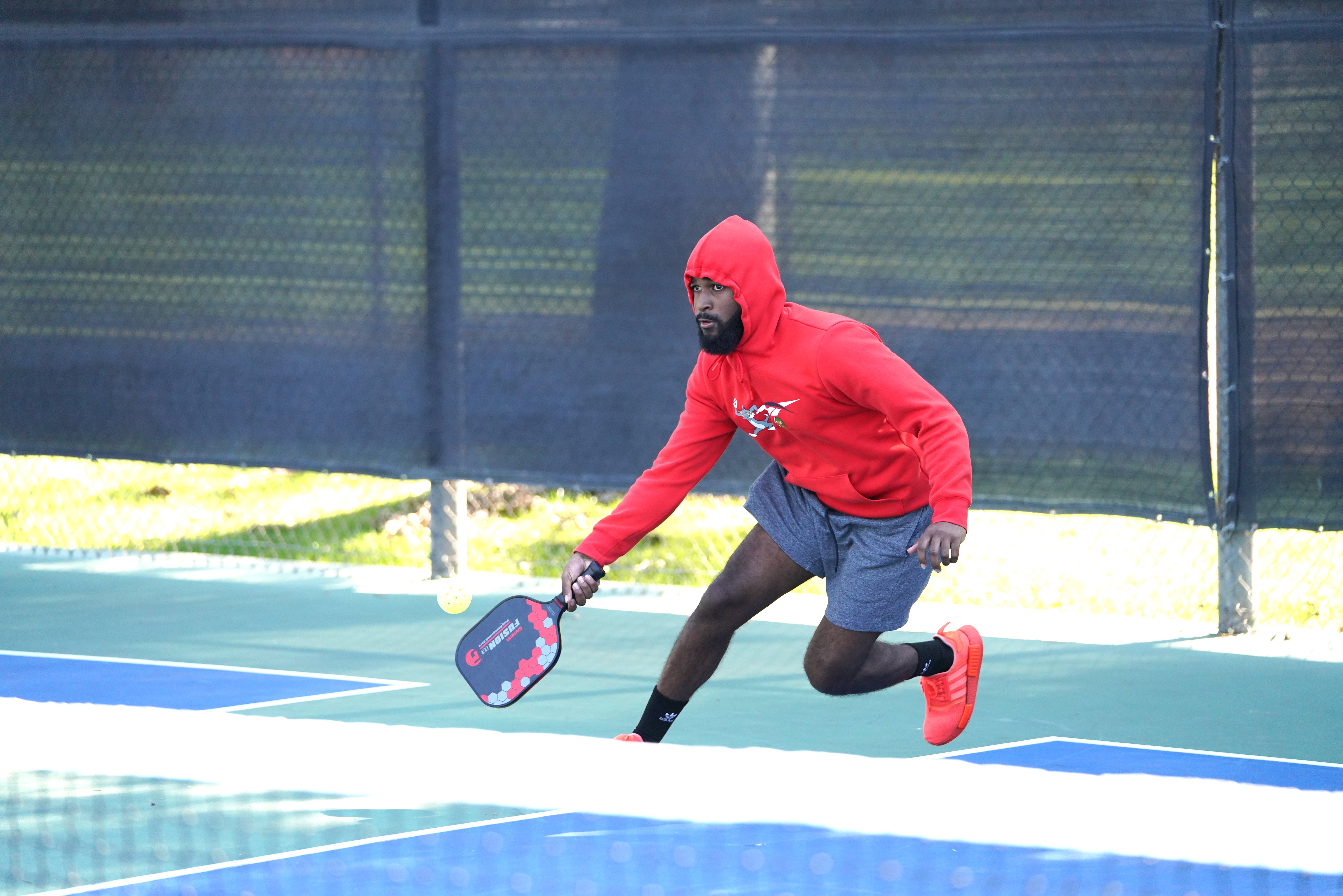 Man in red hoodie and gray shorts lunges to hit a pickleball with a paddle on an outdoor court.