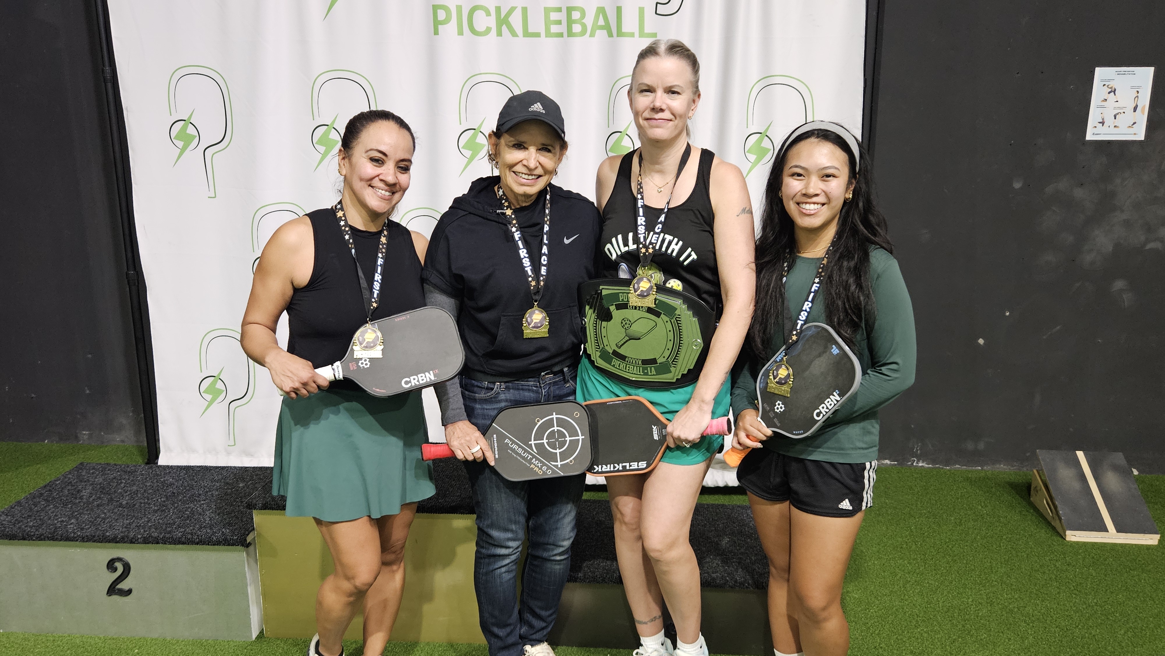 Four women standing on a pickleball podium wearing medals and holding pickleball paddles, with one woman holding a championship belt.
