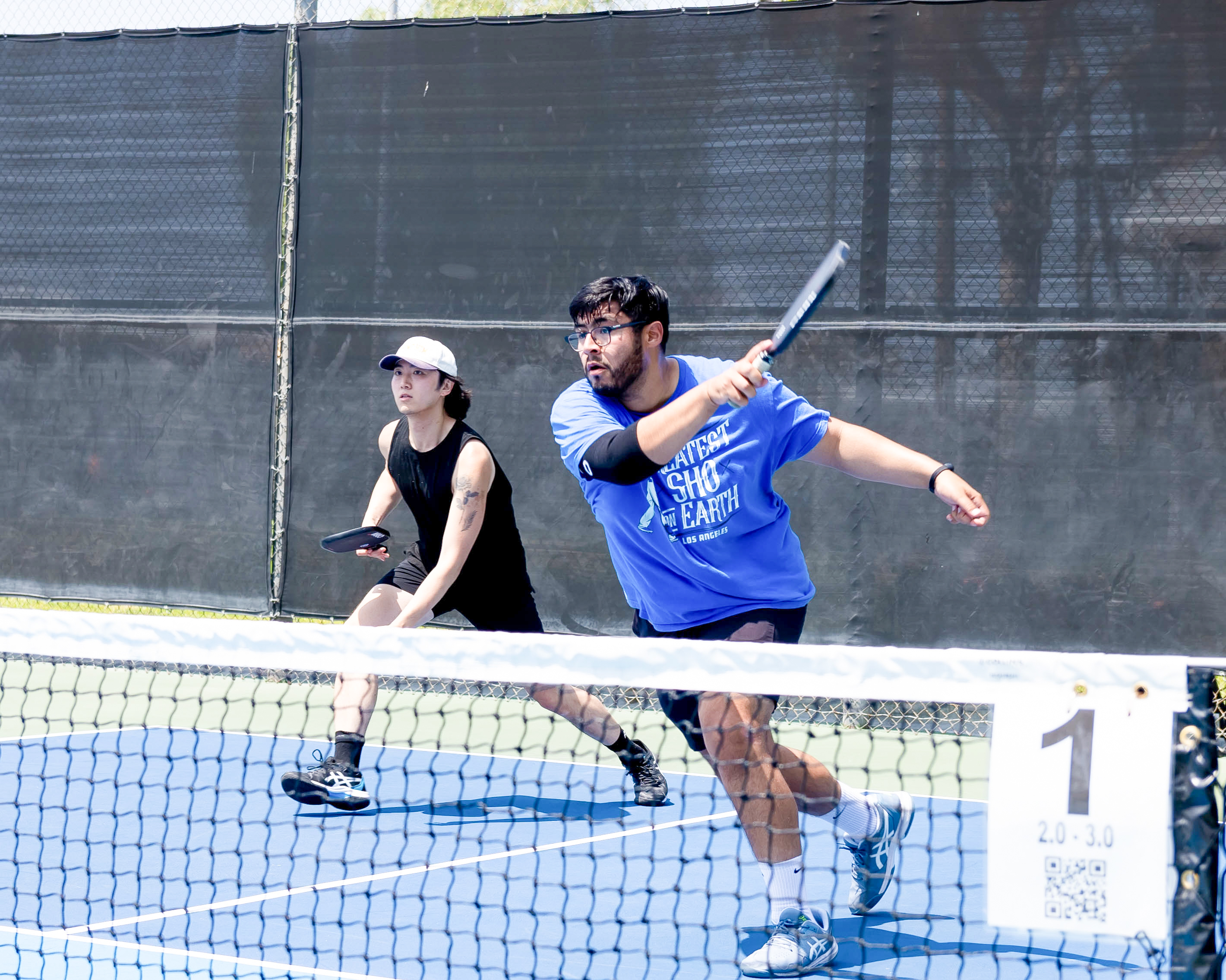 Two men playing pickleball on an outdoor court, one in a blue shirt hitting the ball and the other in black preparing to respond.