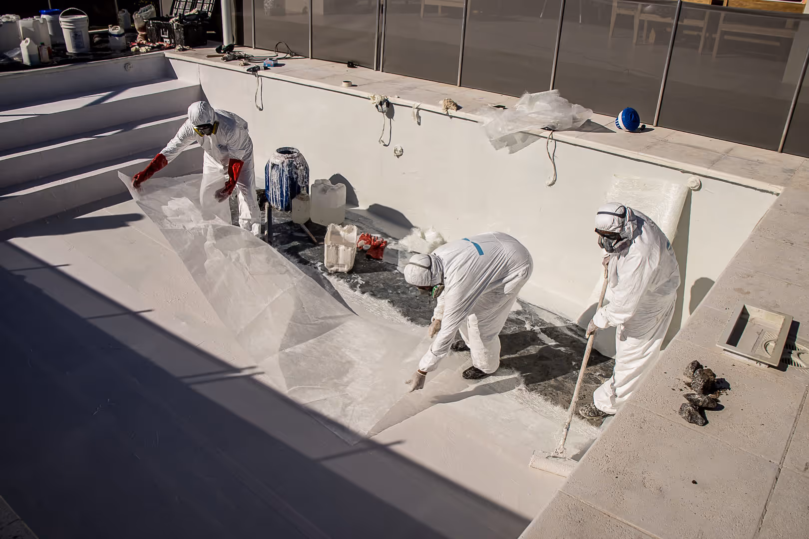 Tres trabajadores con trajes protectores blancos y máscaras aplican una capa blanca dentro de una piscina vacía.