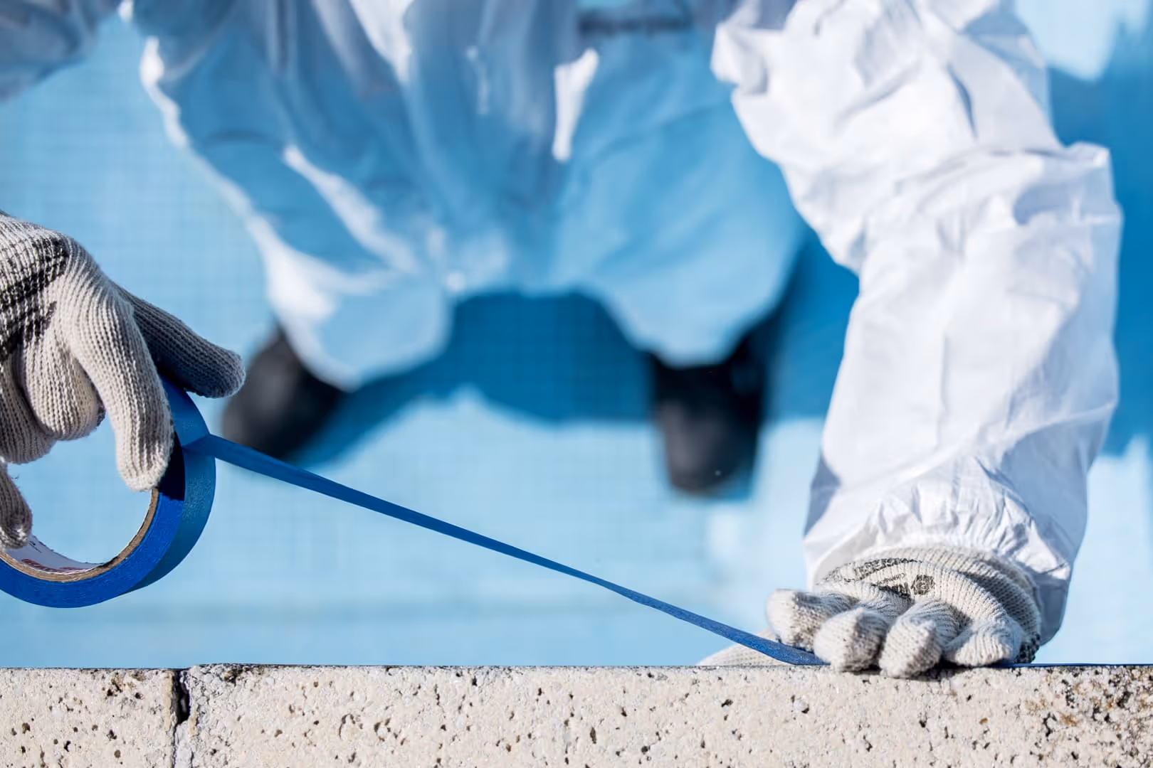 Persona que viste traje protector blanco y guantes tira de cinta de pintor azul en el borde de una piscina.