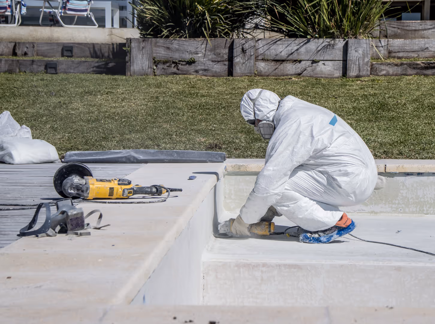 Trabajador con traje de protección y máscara utilizando una herramienta eléctrica para lijar el borde de una piscina vacía.